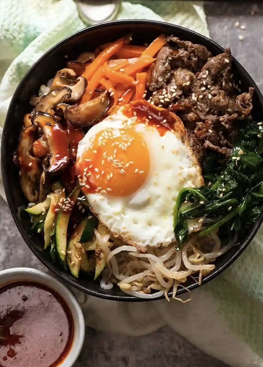 A bowl of bibimbap with beef, assorted vegetables, a fried egg on top, and sauce on the side