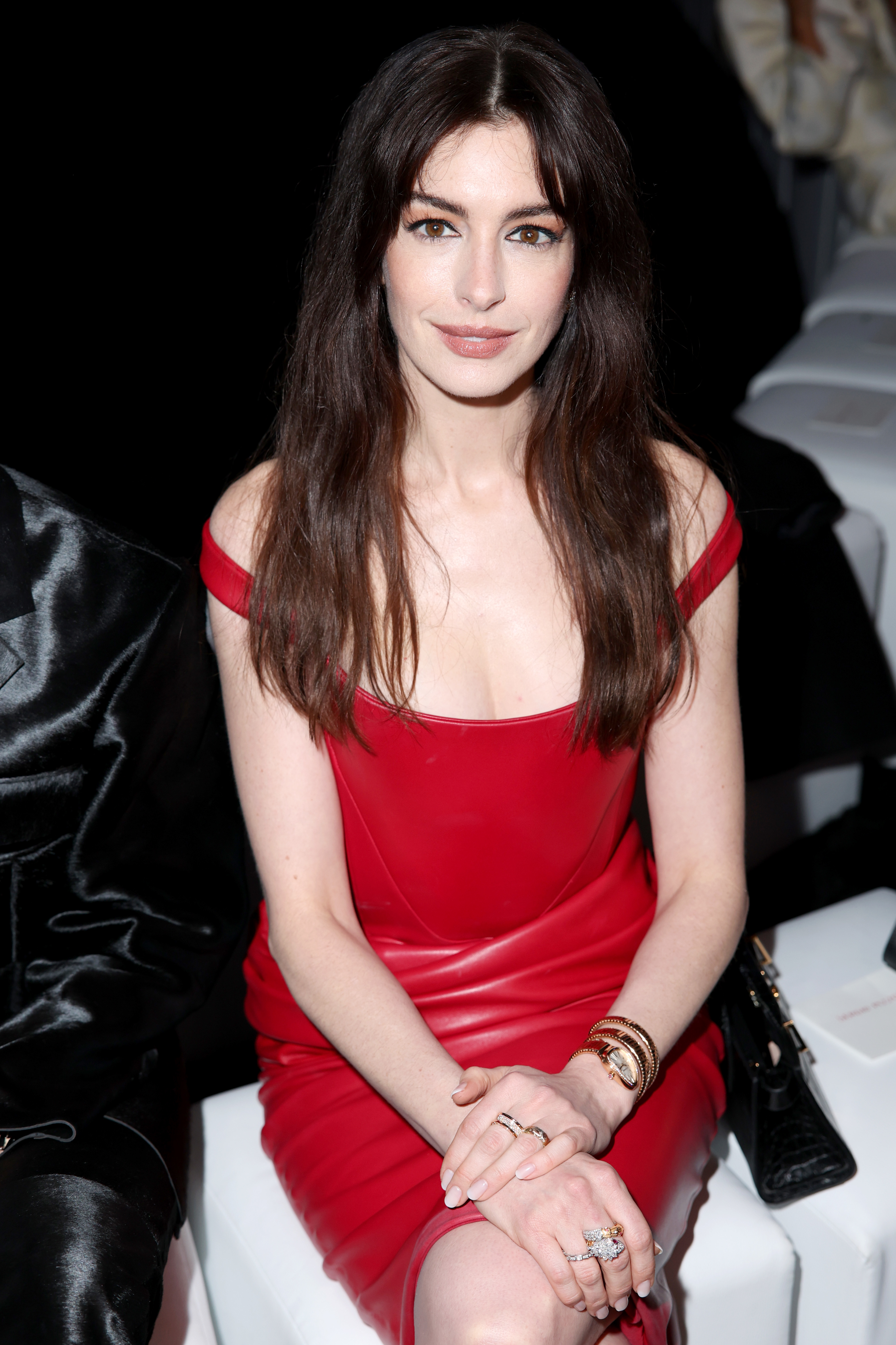 Anne Hathaway in a red off-shoulder dress at an event, seated, with hands clasped