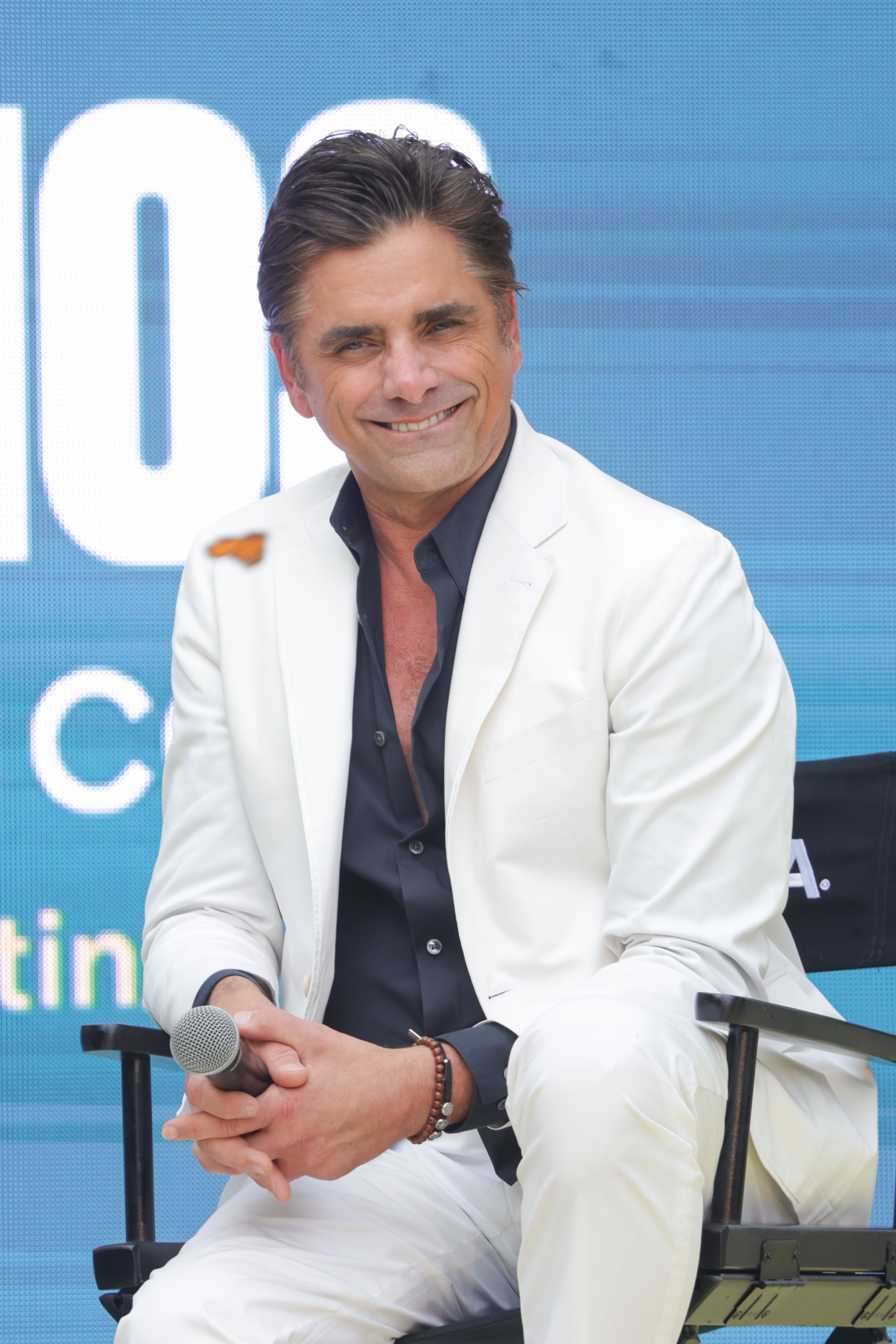 John in a white suit seated with a microphone, smiling during an event