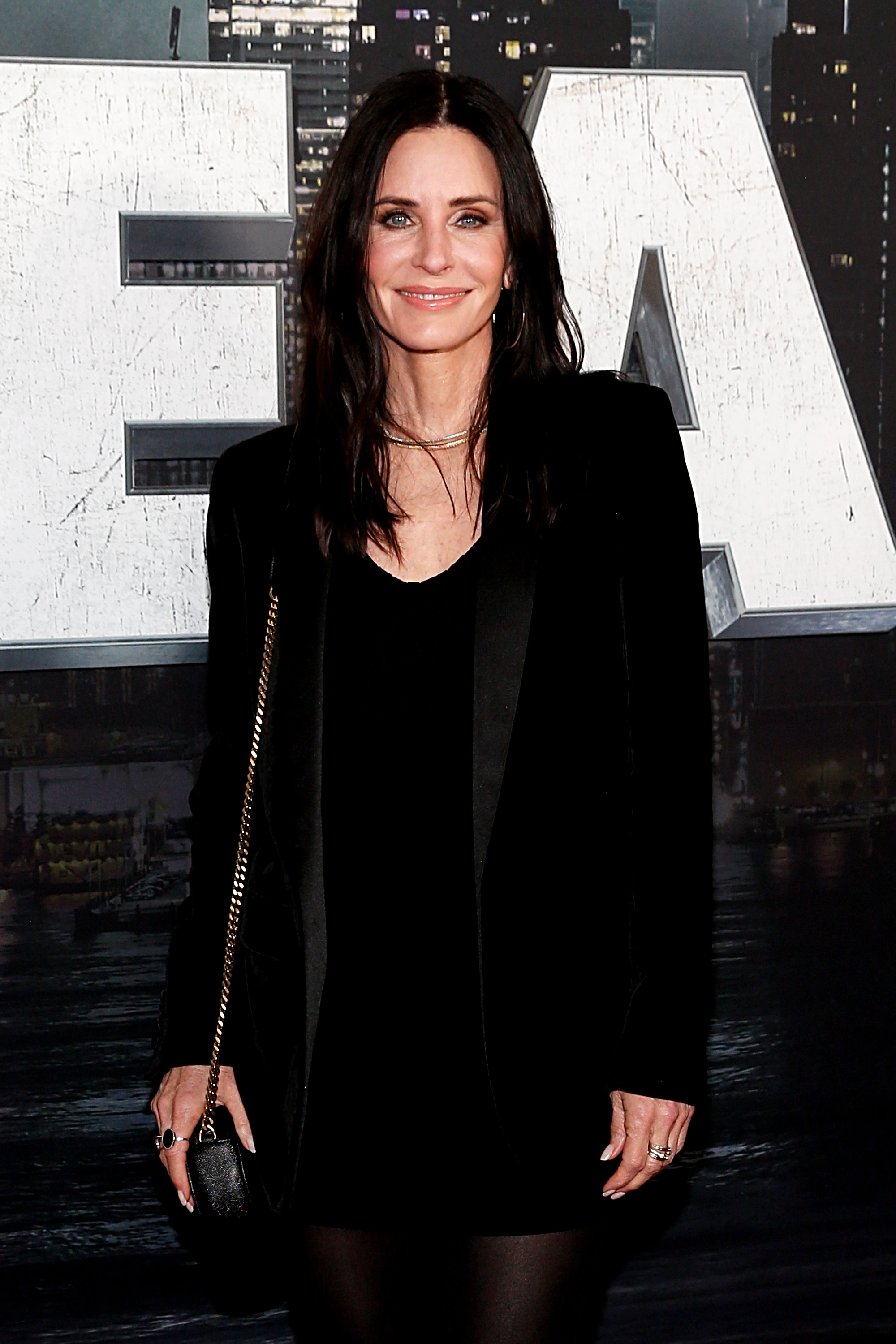 Courteney standing before a &quot;Z&quot; sign, wearing a black blazer over a sheer top, smiling