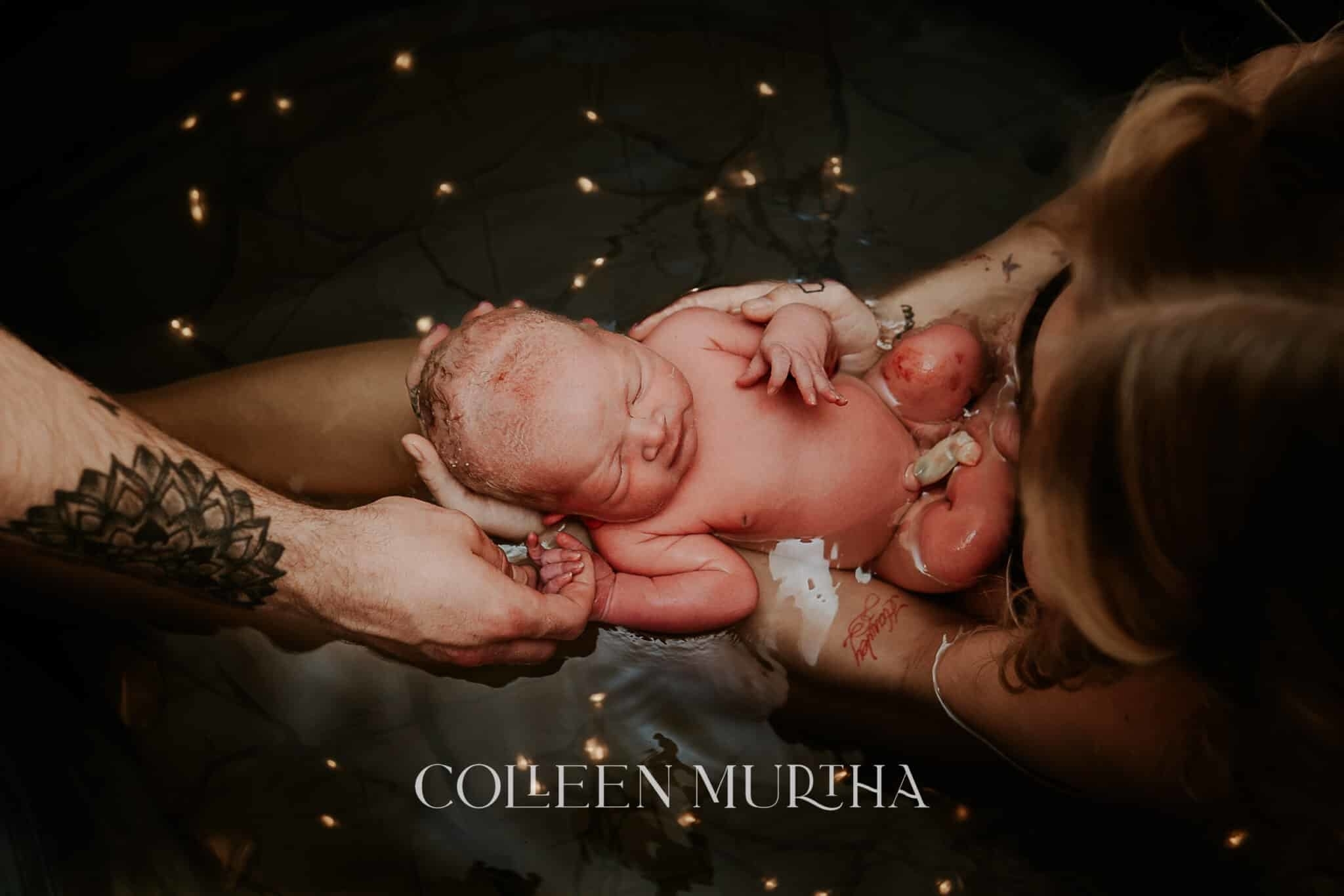 Newborn held by parents' hands in a birthing pool, surrounded by floating candles
