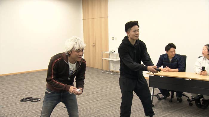 二人の男性が卓上ゲームをしている様子。後ろには座って見守る人が二人いる。