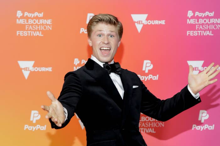 Man in a black tuxedo smiling with arms outstretched at a Melbourne Fashion Festival event