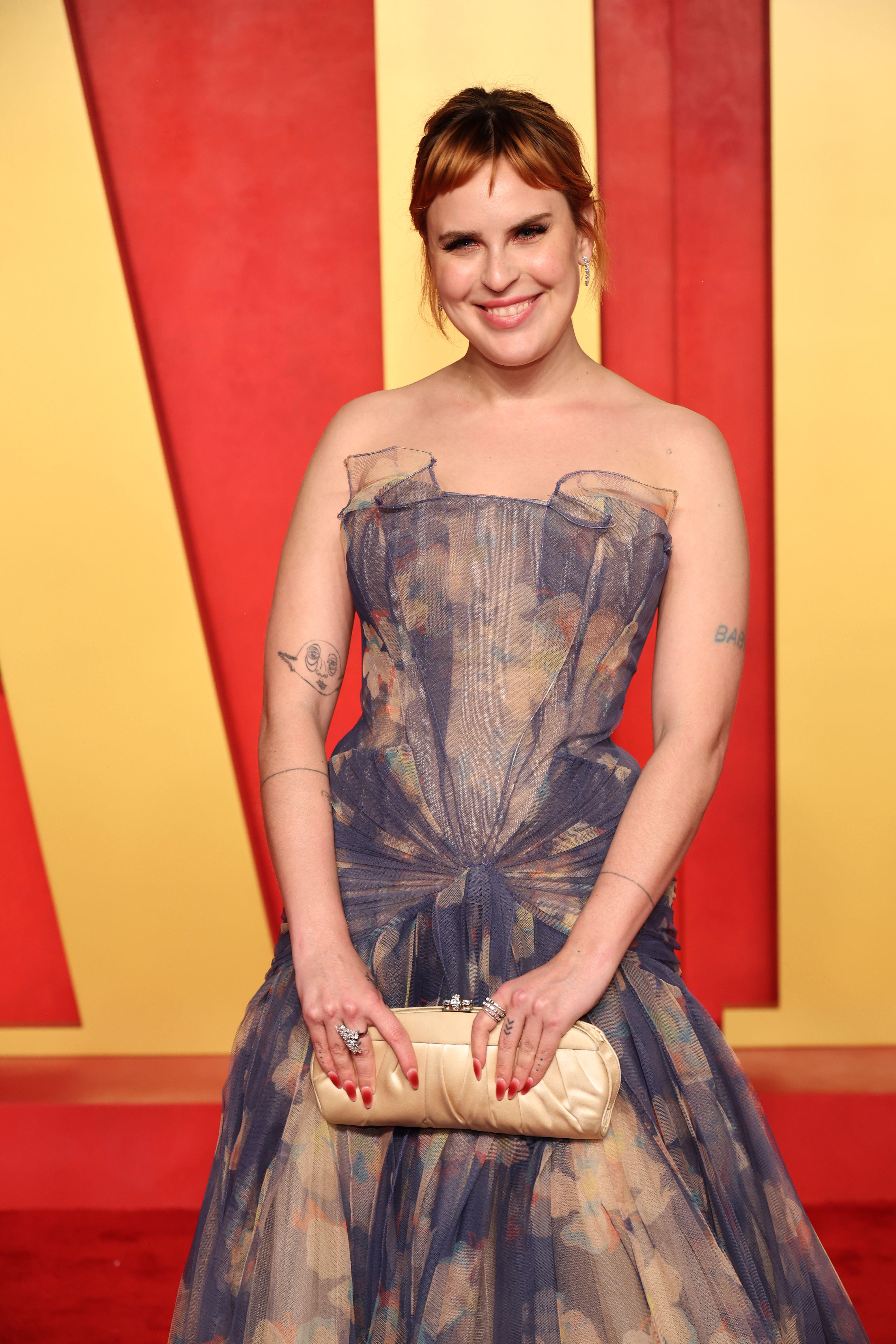 Tallulah smiling in a patterned gown with sheer overlay and bow detail, clutch in hand, standing on a red carpet