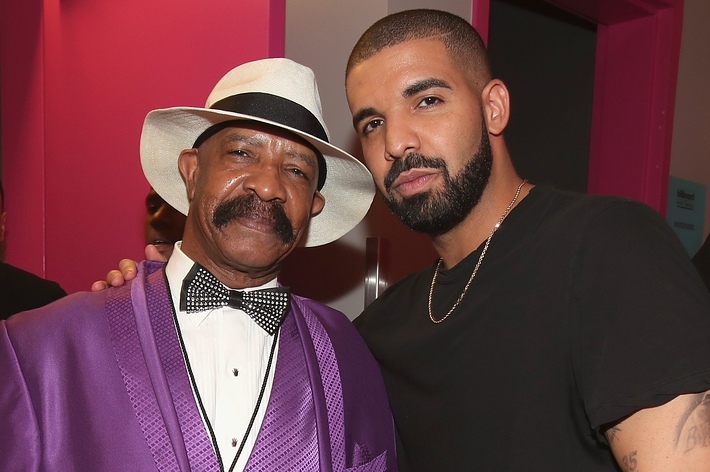 Two men posing for a photo, one in a purple suit and hat with a bow tie, the other in a black short-sleeved shirt
