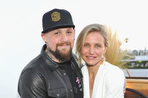 Man in a leather jacket and cap with woman in a white blazer, both smiling, pose together