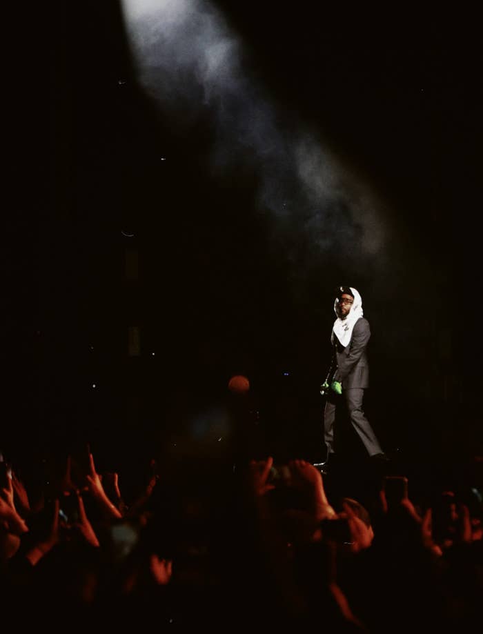 Kendric Lamar en escenario ante multitud con micrófono y guitarra, iluminado por un foco
