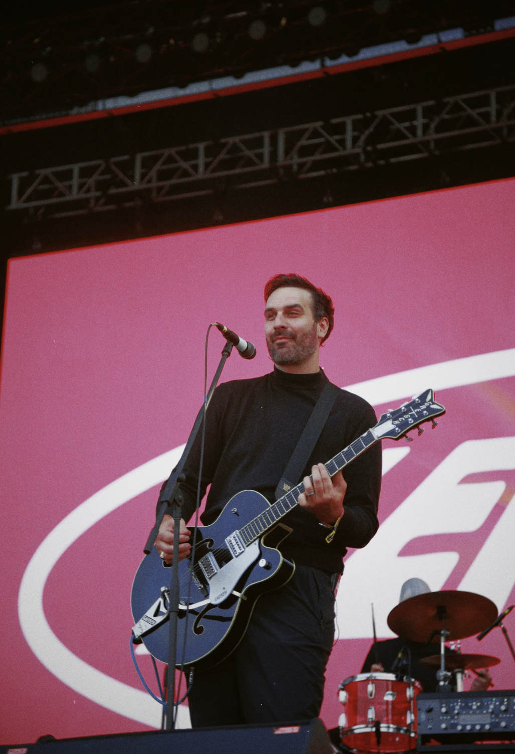 Músico en el escenario tocando una guitarra eléctrica, con una batería detrás y un fondo de pantalla rosa