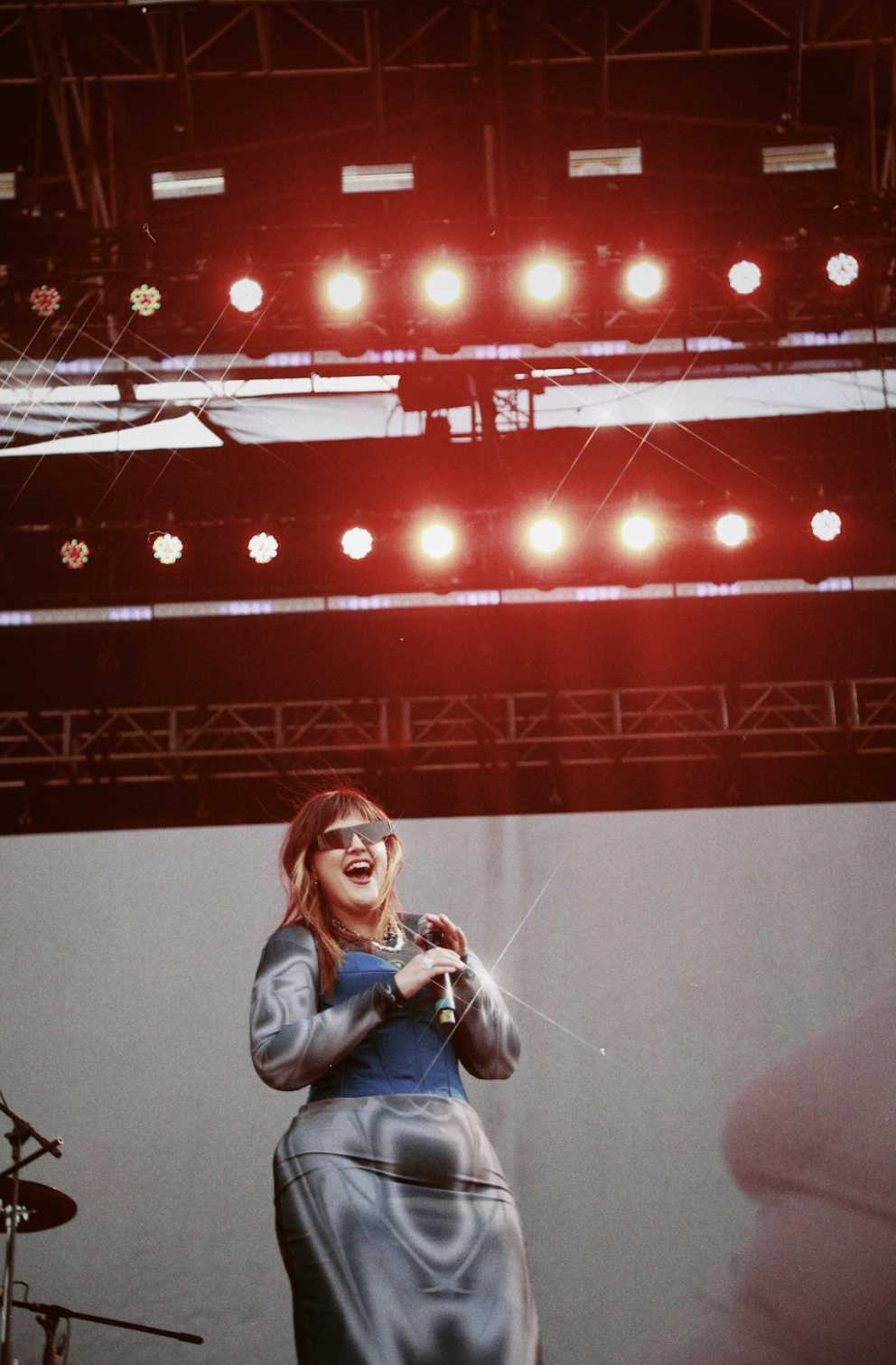 Cantante femenina en el escenario con micrófono, frente a luces brillantes, vestida con traje