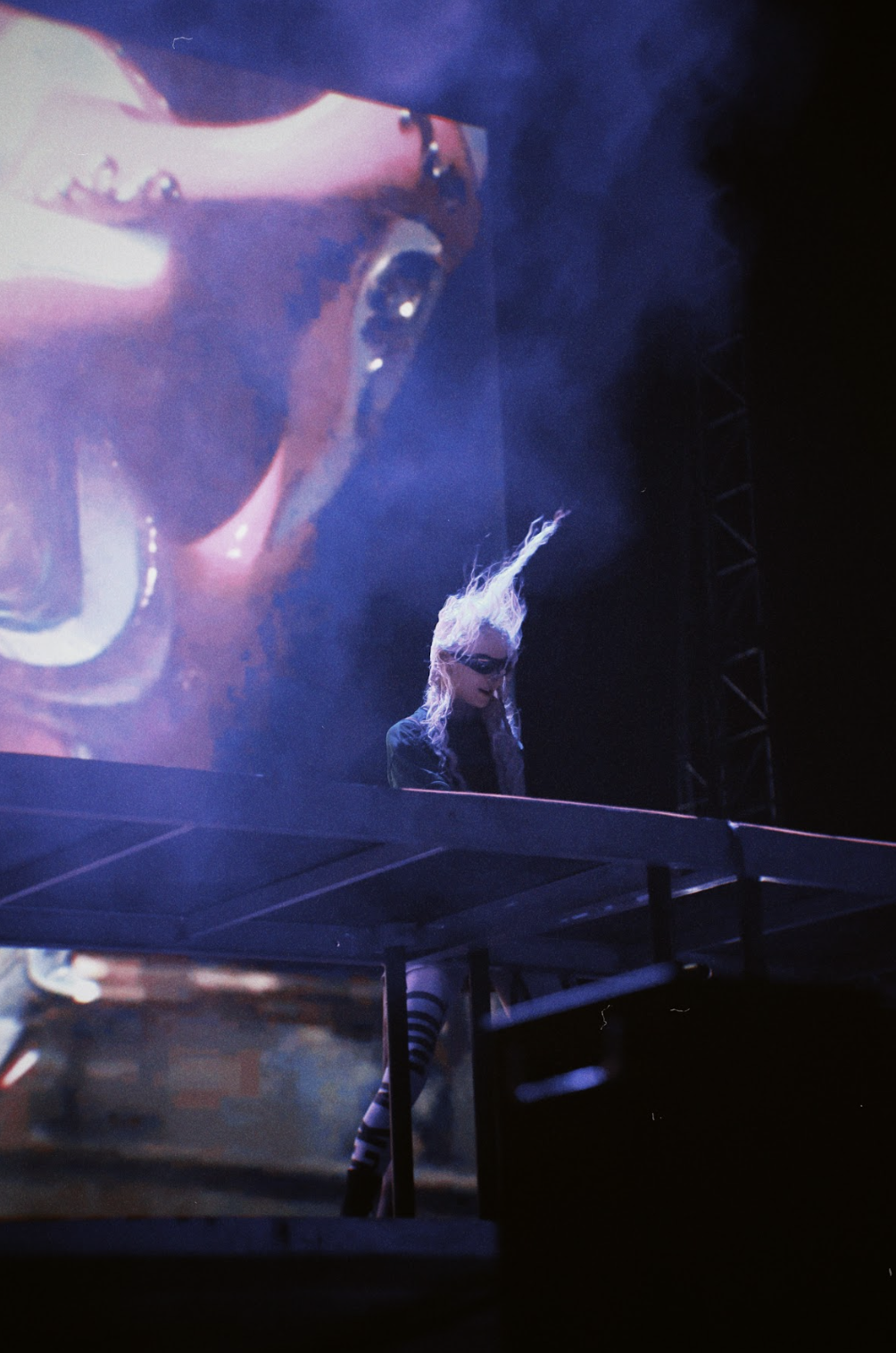 Artista en concierto con vestuario de rayas y tocado extravagante, frente a pantalla gigante