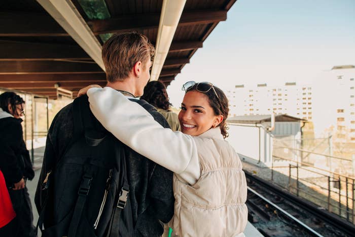 Two people embracing at a train station, the man facing away, the woman smiling at the camera behind her