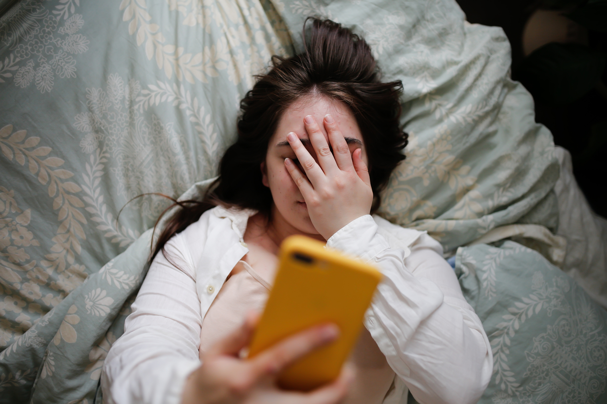 Person in bed holding phone with hand on face, looks stressed or tired