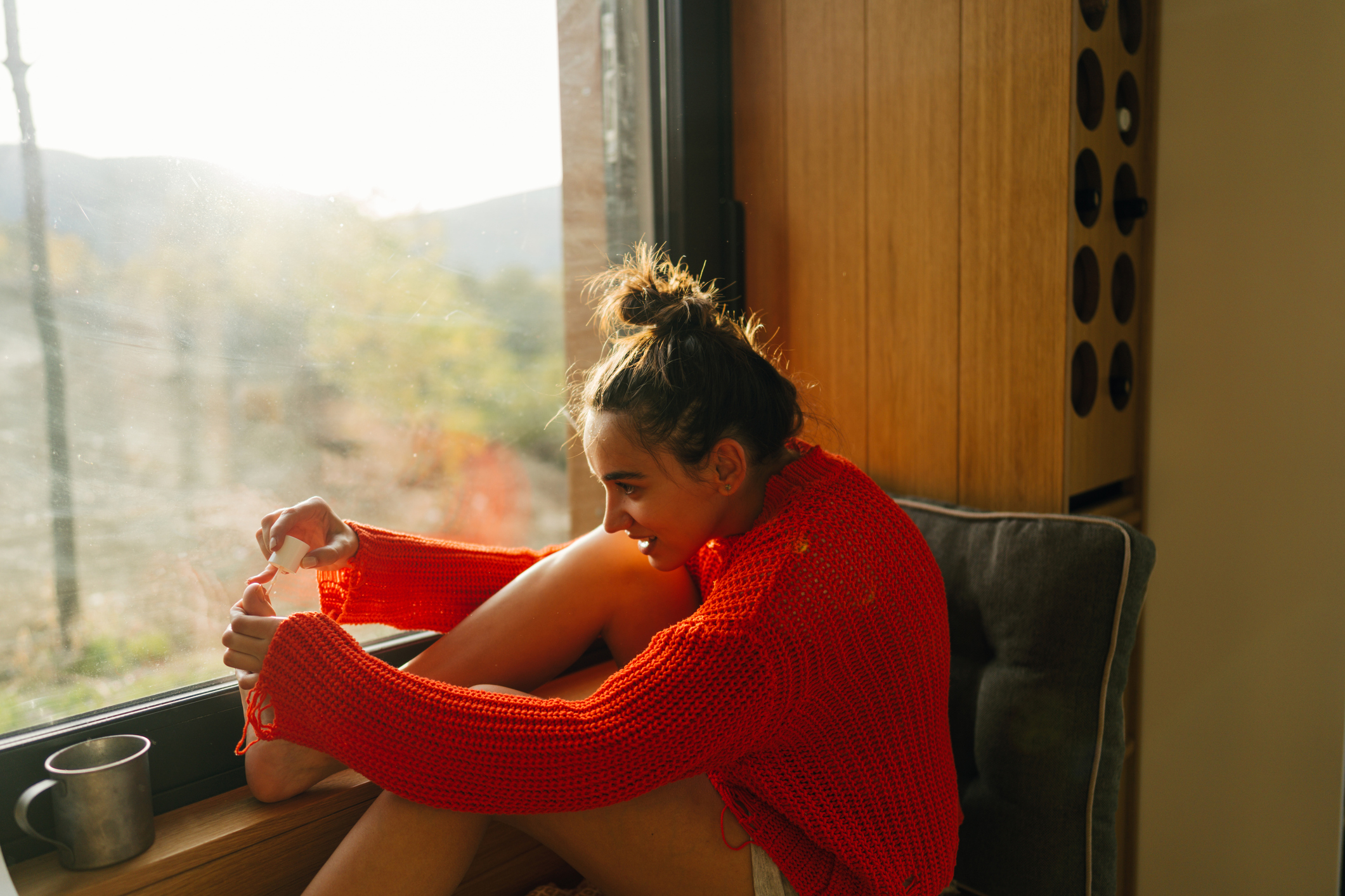 Woman seated by window, smiling, in cozy red knitwear, applying toenail polish