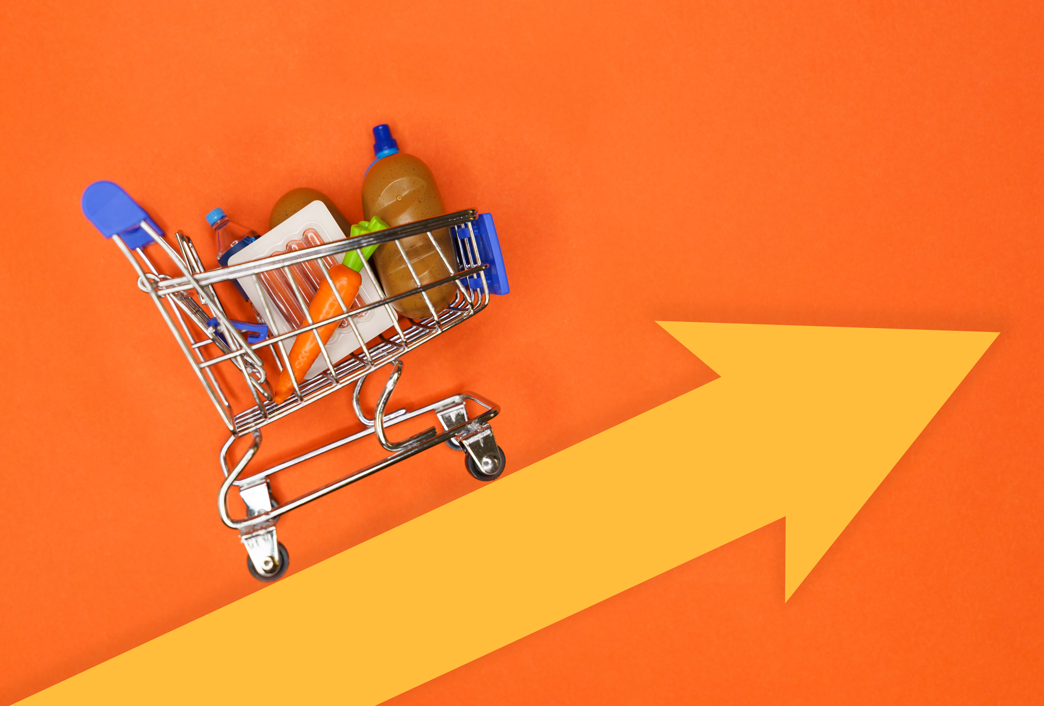 Mini shopping cart filled with groceries on orange background with a large yellow arrow pointing right