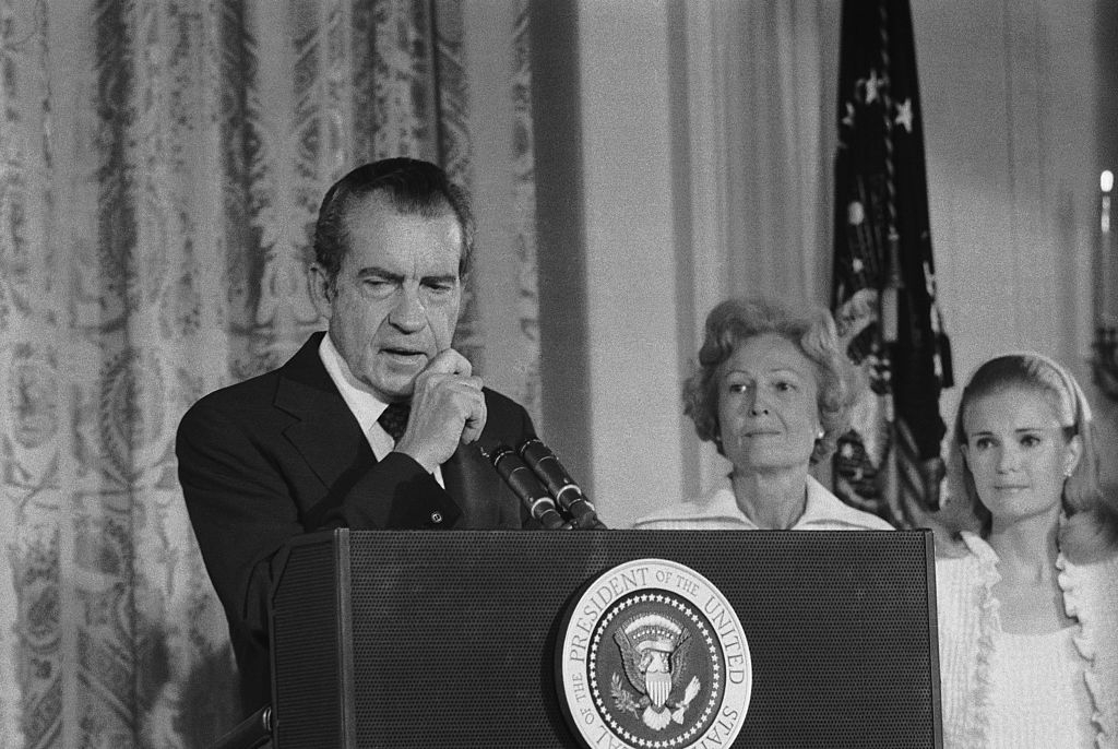 Man in suit stands at a podium with presidential seal, thoughtful expression, others present