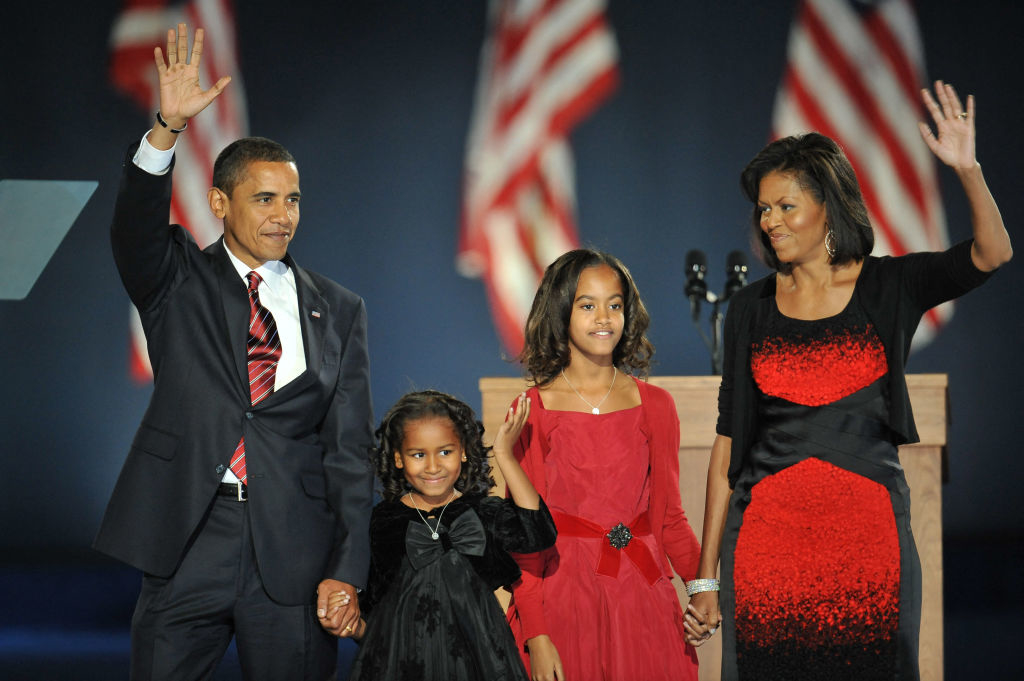 Barack Obama with daughters and Michelle Obama on stage, waving to the crowd, dressed in formal attire