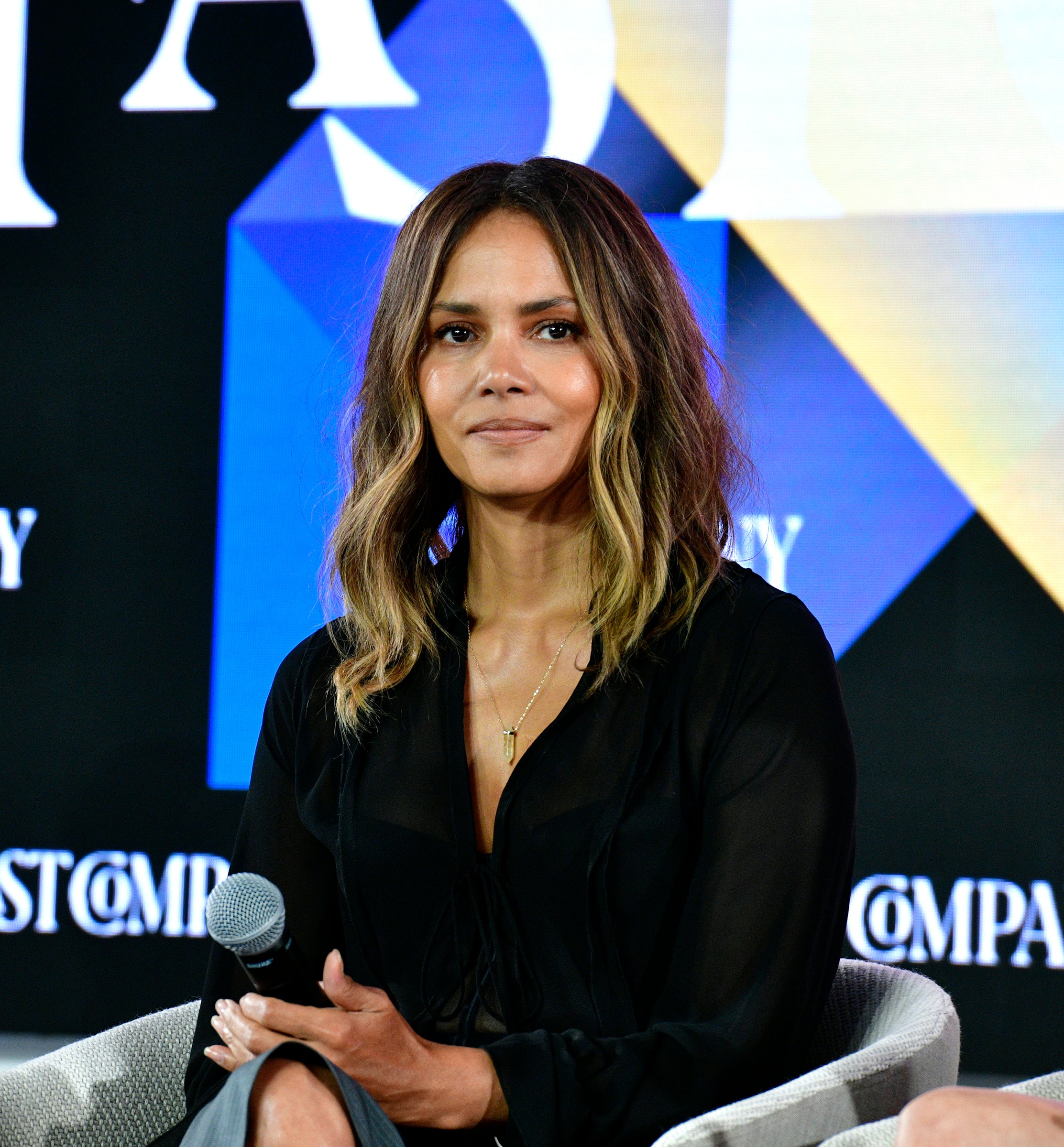 Halle Berry sits, speaking at a Fast Company event, wearing a black v-neck dress with her legs crossed