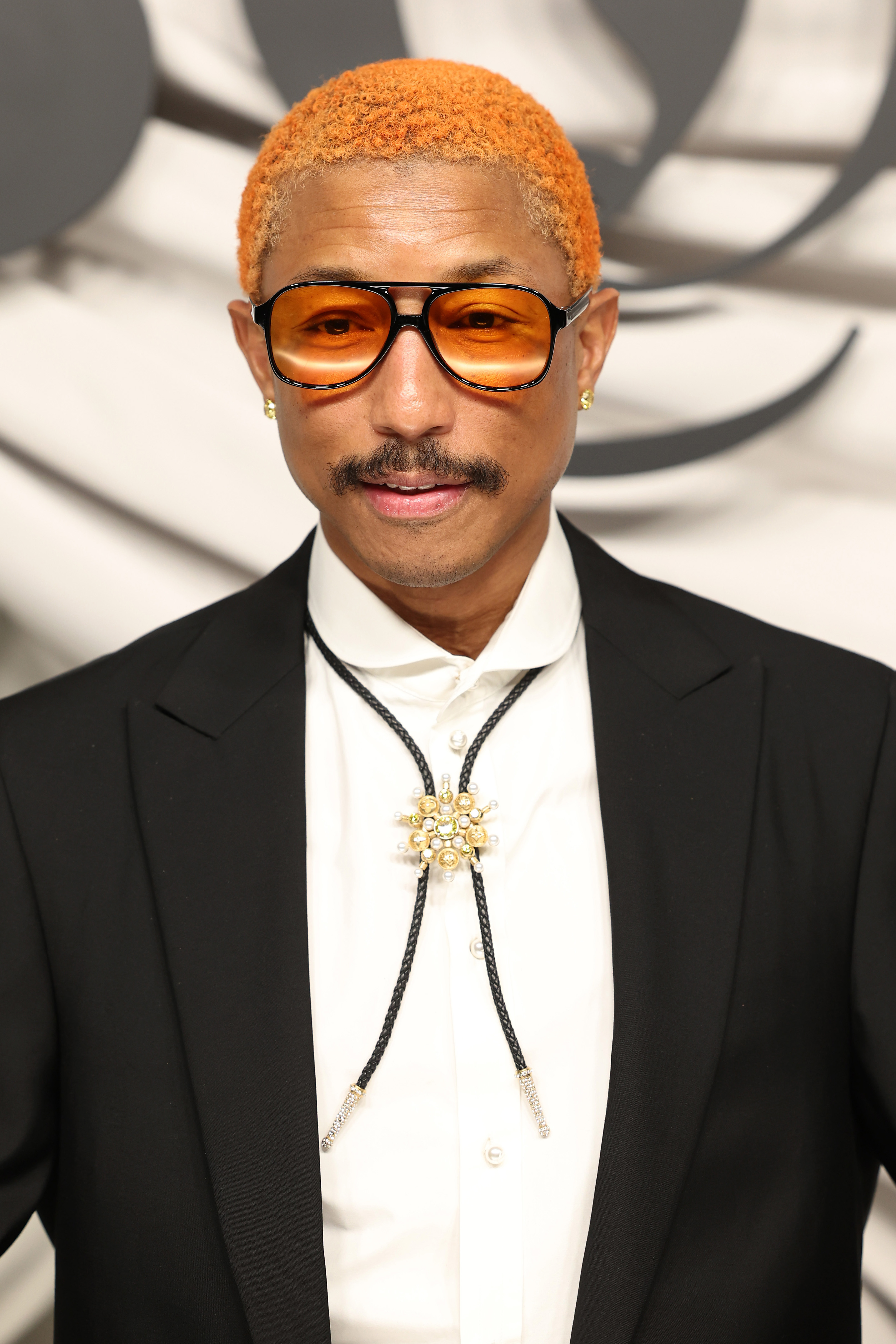 Pharrell with orange  hair and large glasses, wearing a black suit and ornate brooch on a white shirt