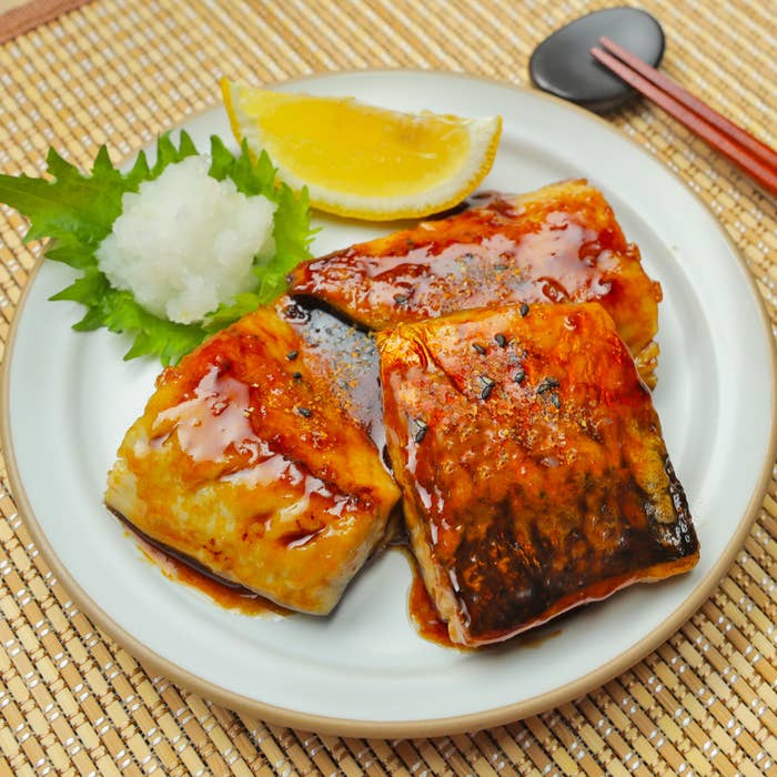 焼き魚にタレをかけ、添え物に大根おろしとレモンスライスがある