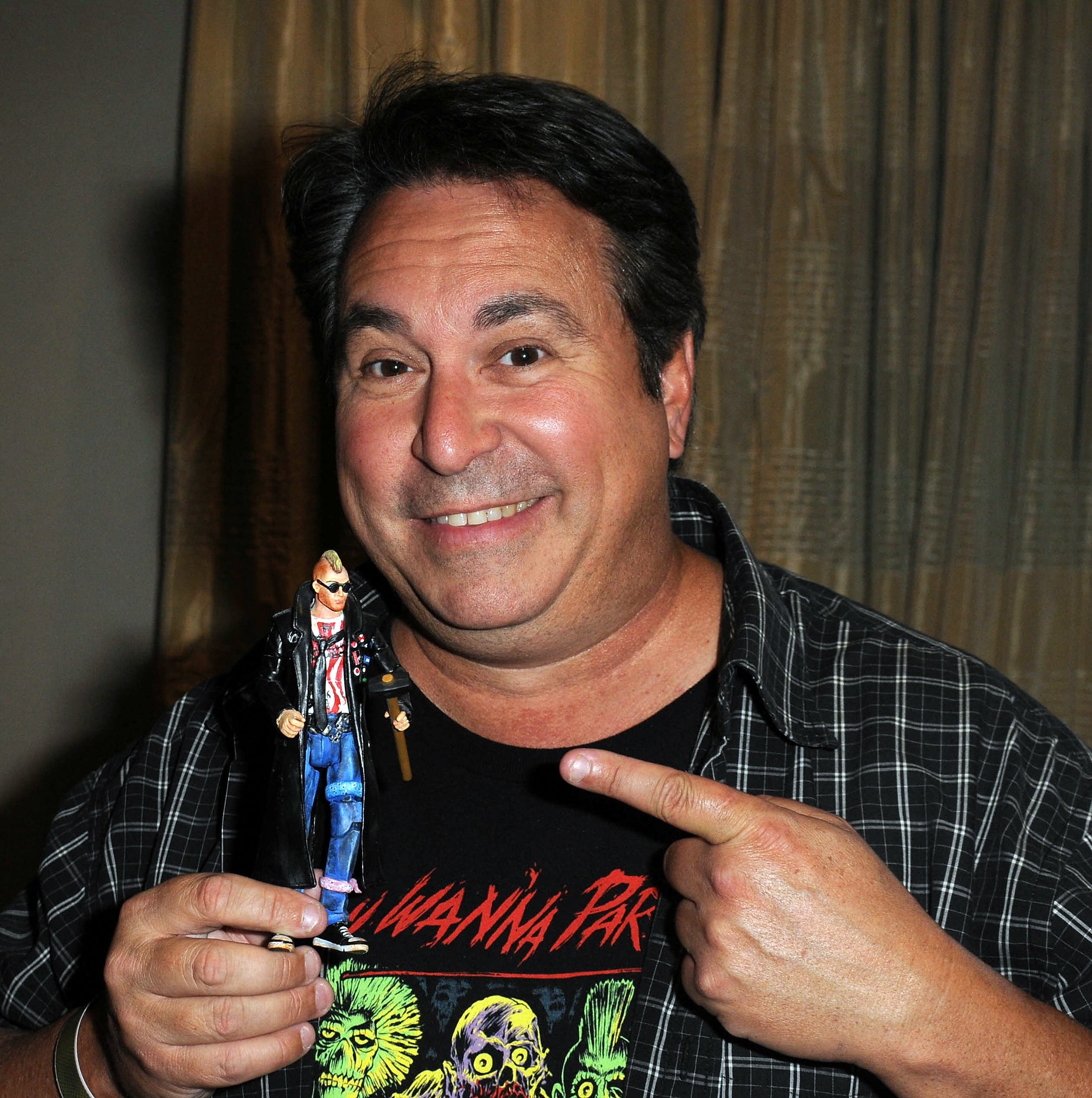 Brain smiling, holding and pointing to a small figurine, wearing a graphic t-shirt with plaid over-shirt