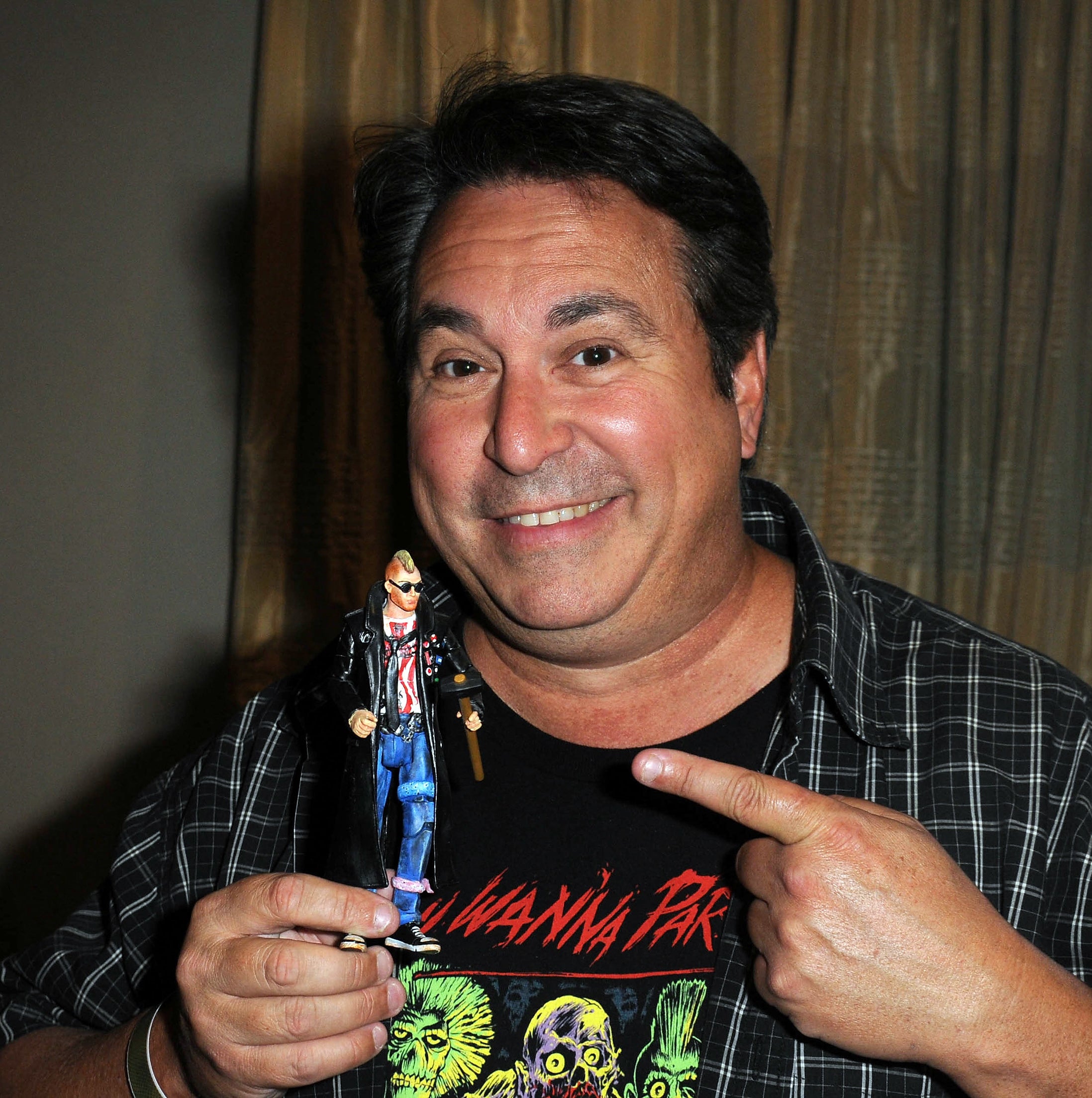 Man smiling, holding and pointing to a small figurine, wearing a graphic t-shirt with plaid over-shirt