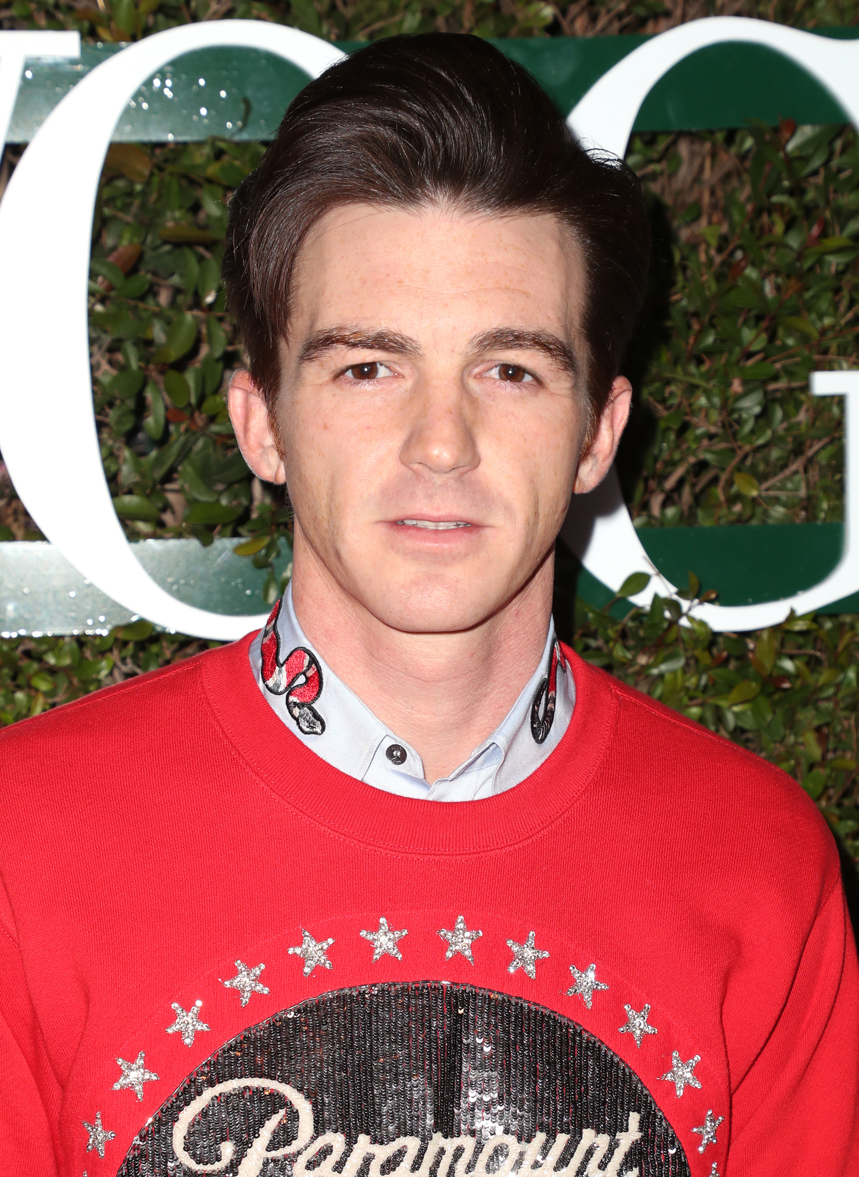 Drake Bell wearing a red sweater with Paramount logo, posing in front of a hedge backdrop