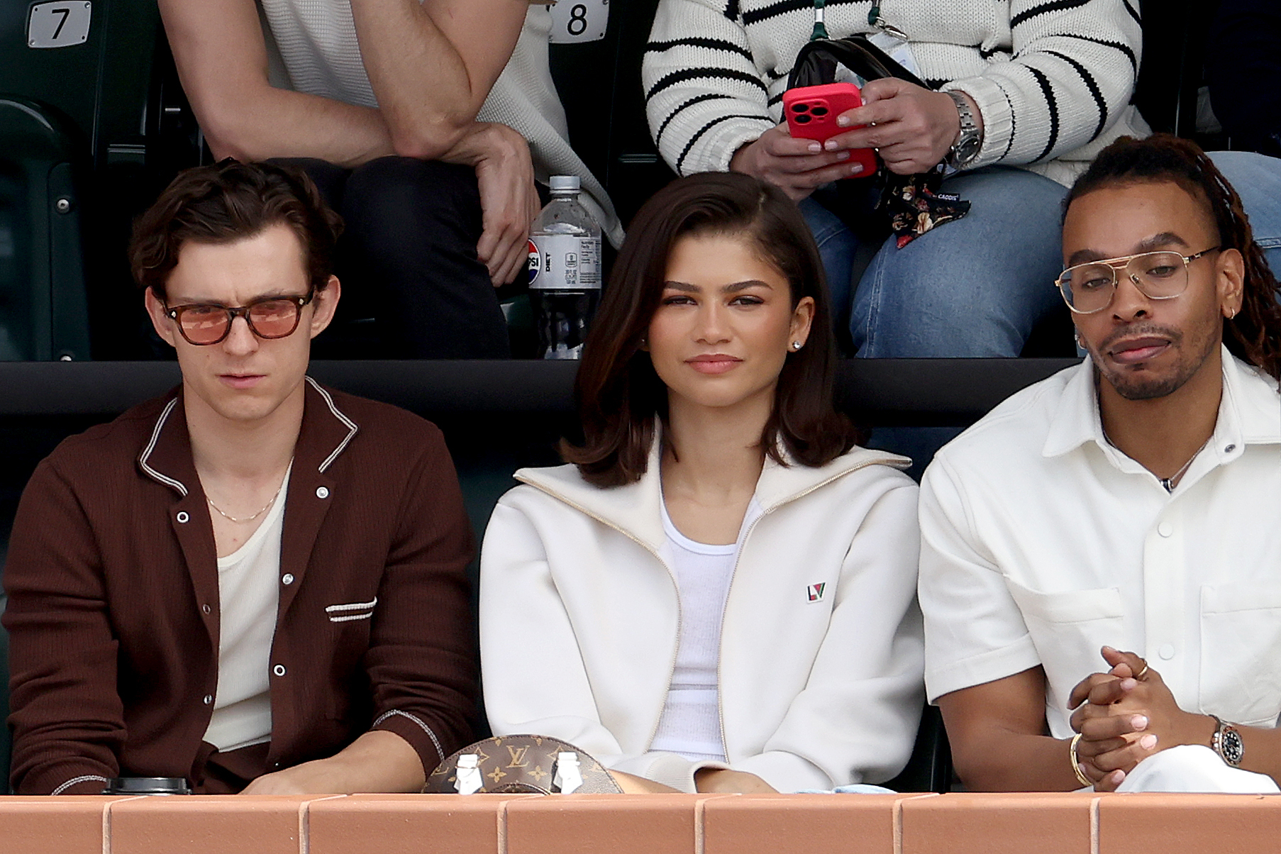 Tom Holland, Zendaya, and a third person sit side by side at an outdoor event, appearing attentive