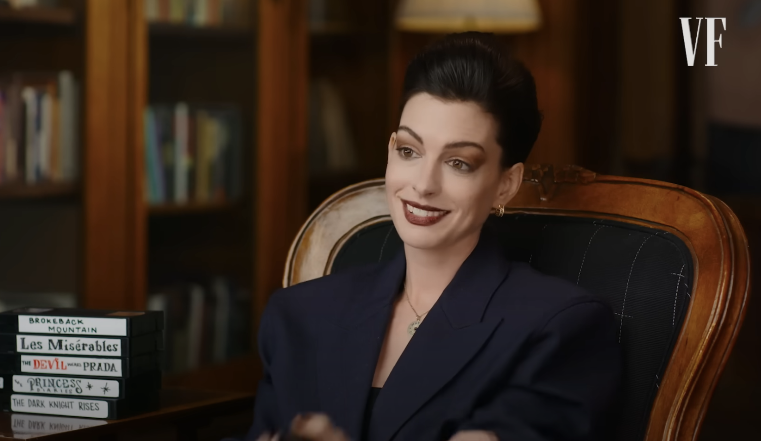 Anne Hathaway in a dark blazer, seated, gesturing mid-interview with books in the background
