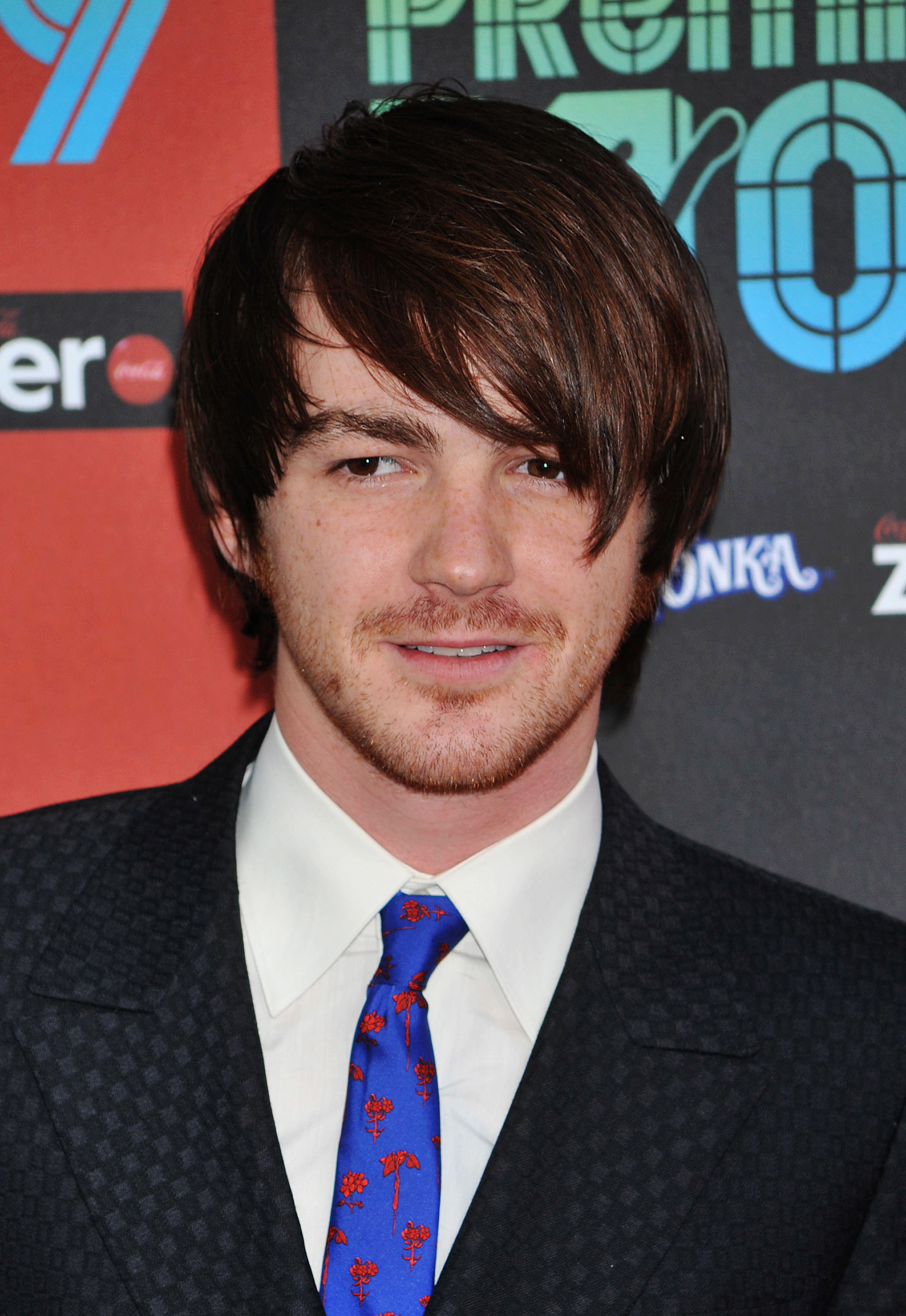 Drake Bell in a suit with a patterned tie, at an event with logos in the background