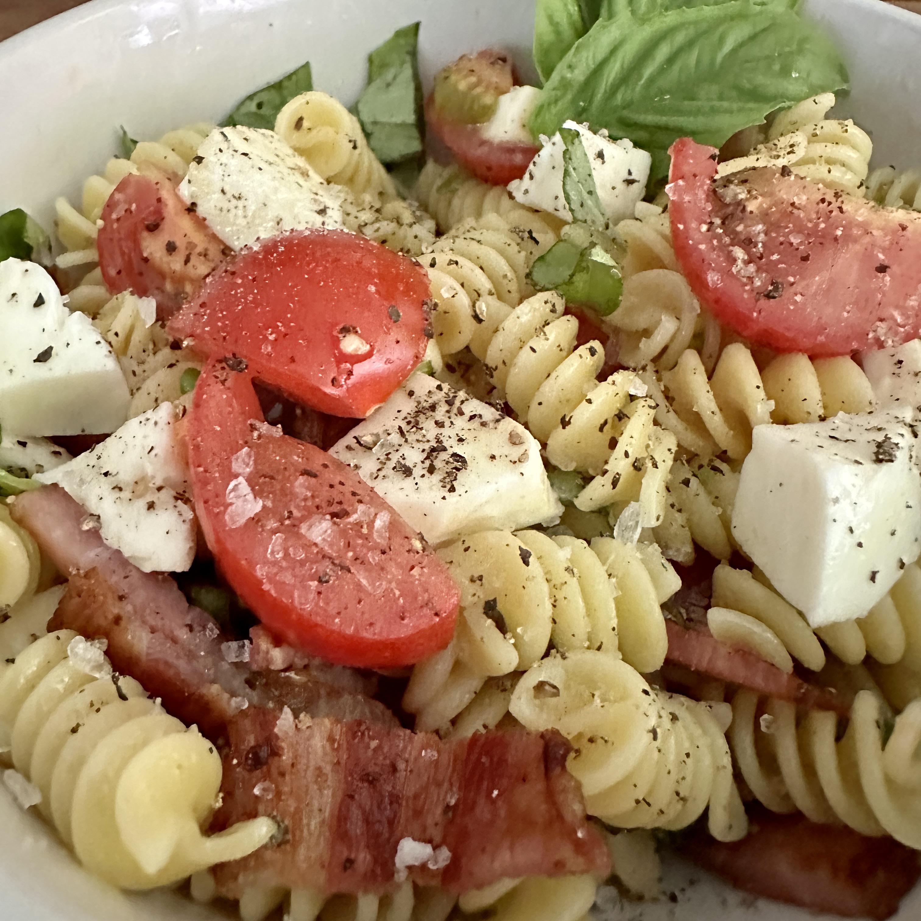 Bowl of pasta salad with tomatoes, mozzarella, basil, and bacon seasoned with pepper