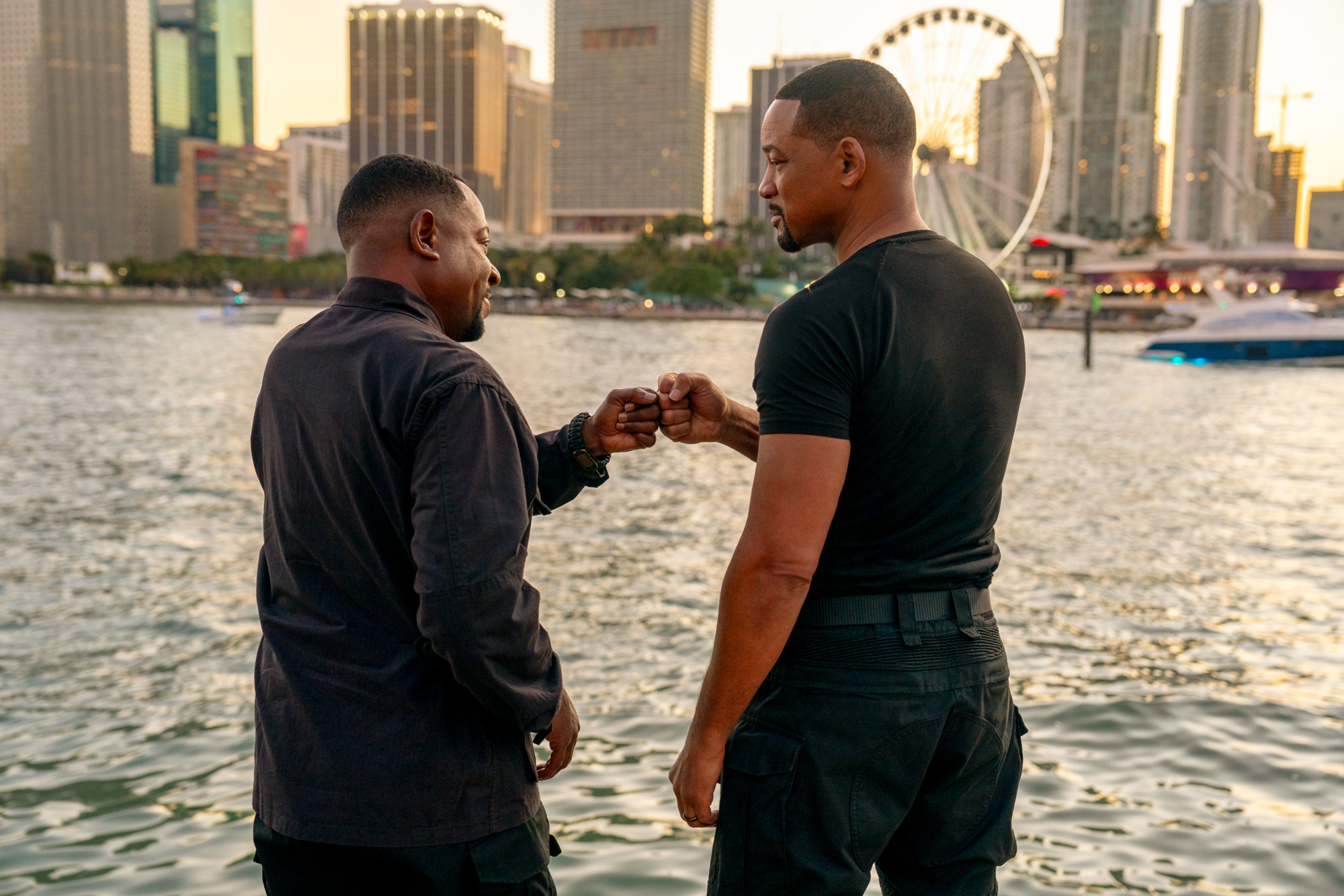 Two actors on set, one touching the other&#x27;s shoulder, with a cityscape and ferris wheel in the background