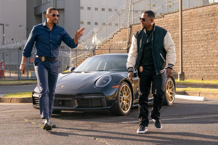 Two men walking and talking beside a Porsche, one in casual attire and the other in a jacket with gold details