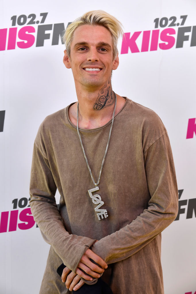 Aaron standing in front of promotional backdrop, wearing a necklace with &quot;LOVE&quot; pendant and casual attire