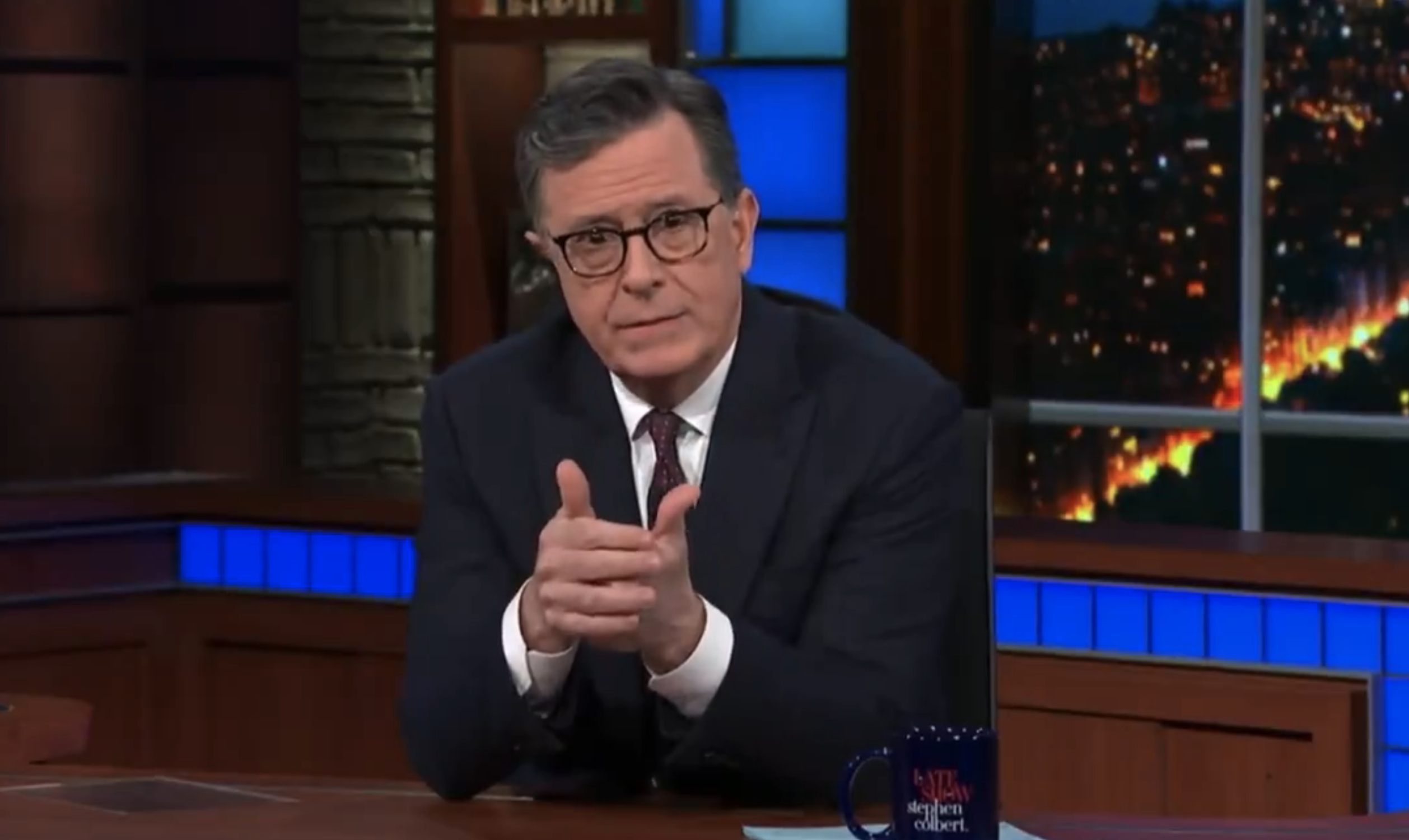 Stephen Colbert, wearing a suit, seated with hands together on his late-night show set
