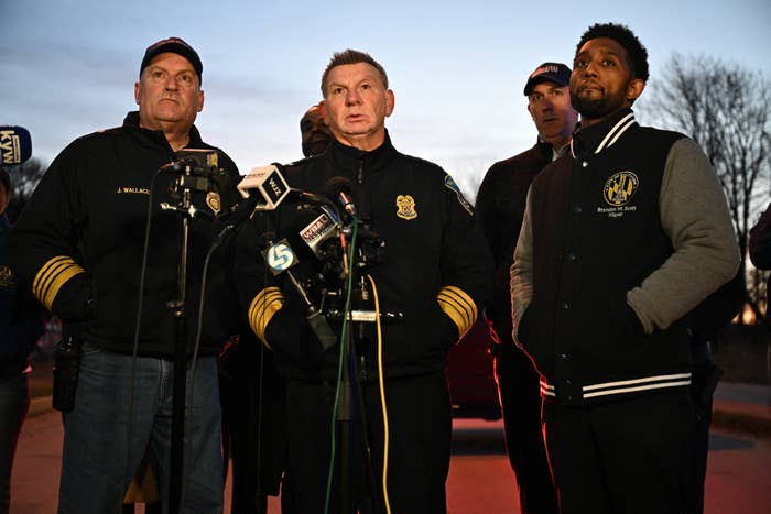 Officials in uniforms and a man in a black jacket at a press conference with microphones