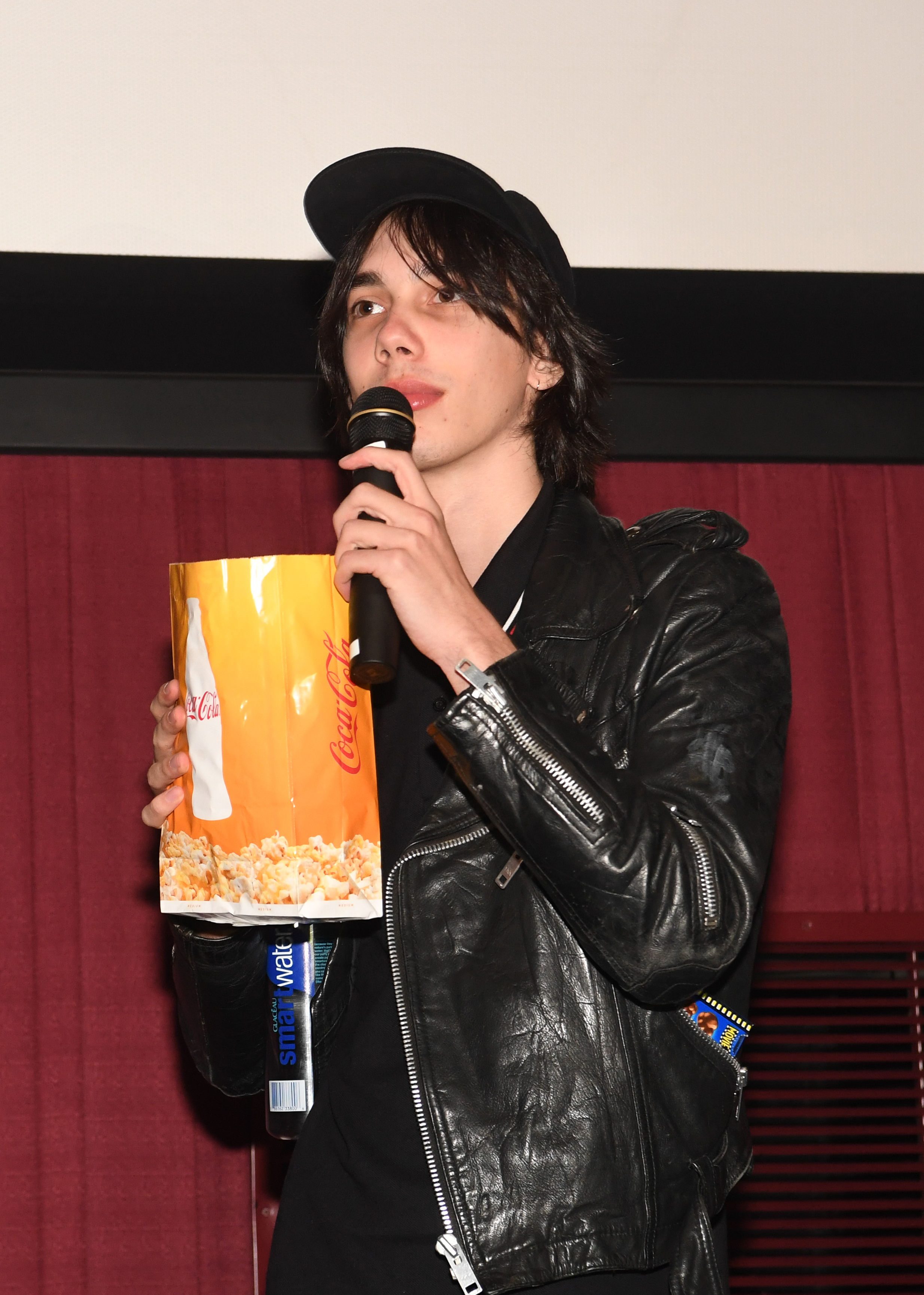 Charlie in a leather jacket and cap holding a microphone and popcorn bucket