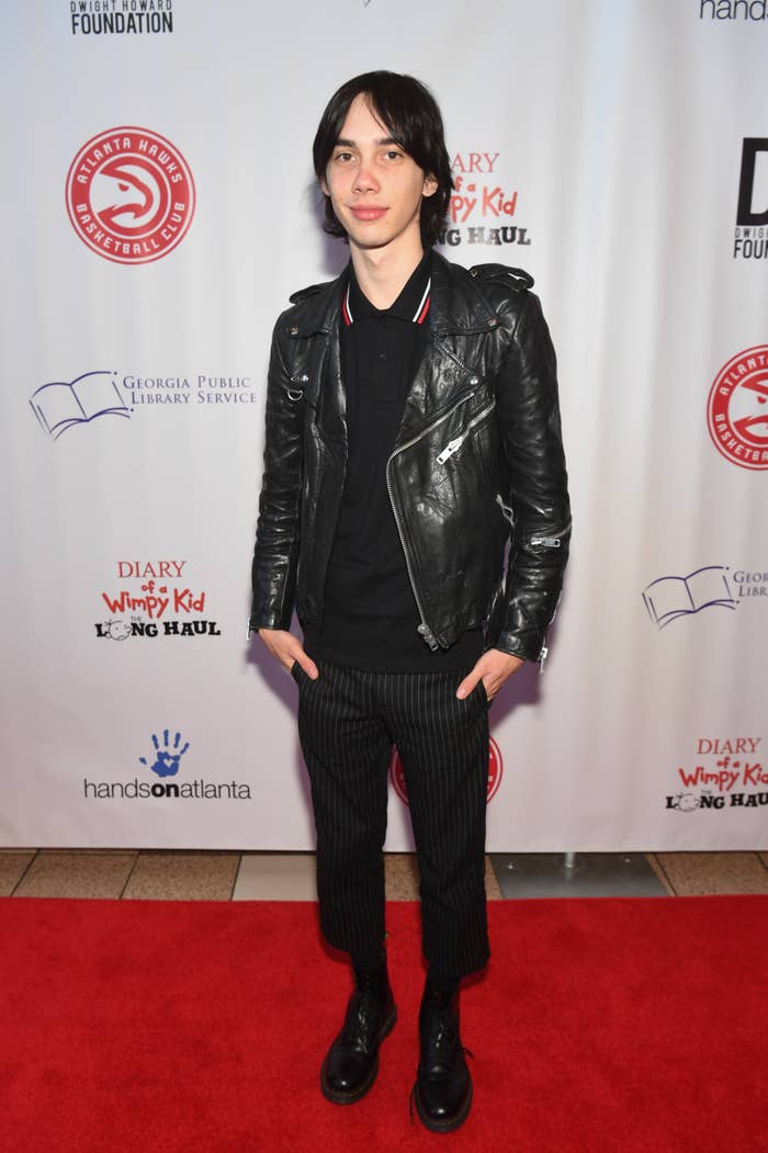 Charlie standing on red carpet wearing a black leather jacket, striped pants, and black shoes, hands in pockets