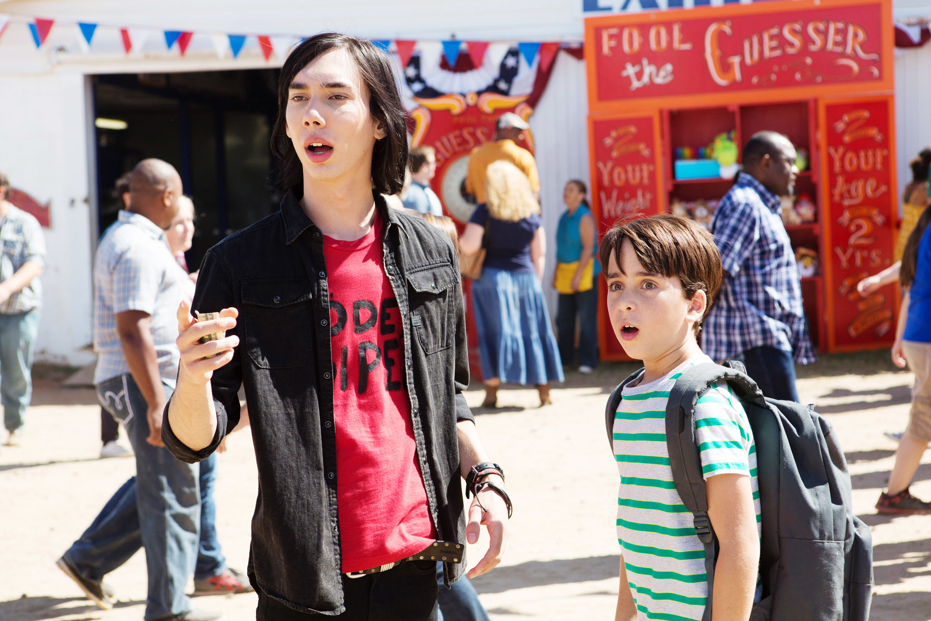 Two kids from &quot;Diary of a Wimpy Kid&quot; stand at a fairgrounds, one eating a hot dog, the other carrying a backpack