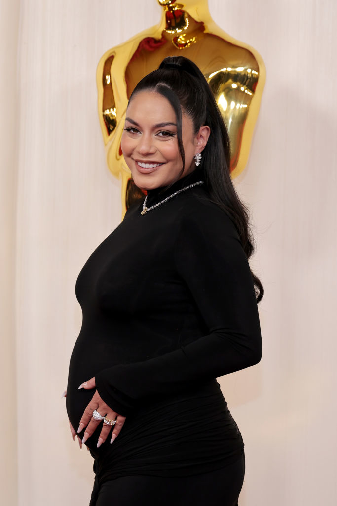 Pregnant Vanessa in a black dress poses with a hand on her bump and smiling
