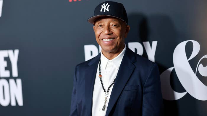 Russell Simmons wearing a blazer and baseball cap with a beaded necklace, smiling at an event
