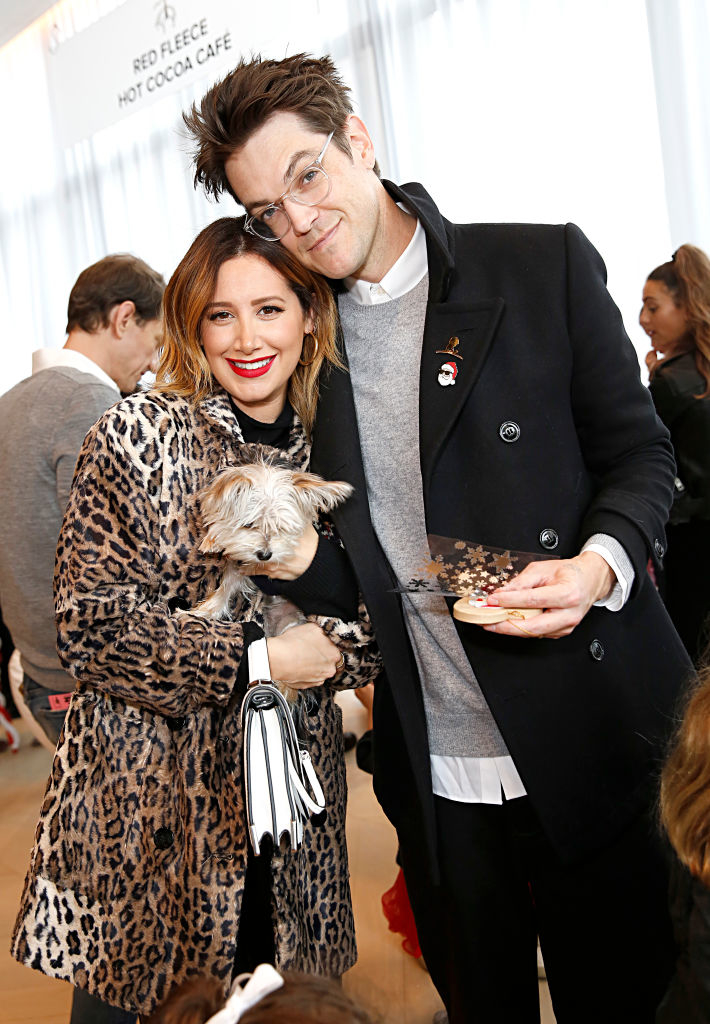 Ashley and her husband at an event, she holding a small dog and clutch, he holding a plate; both are smiling and casually dressed