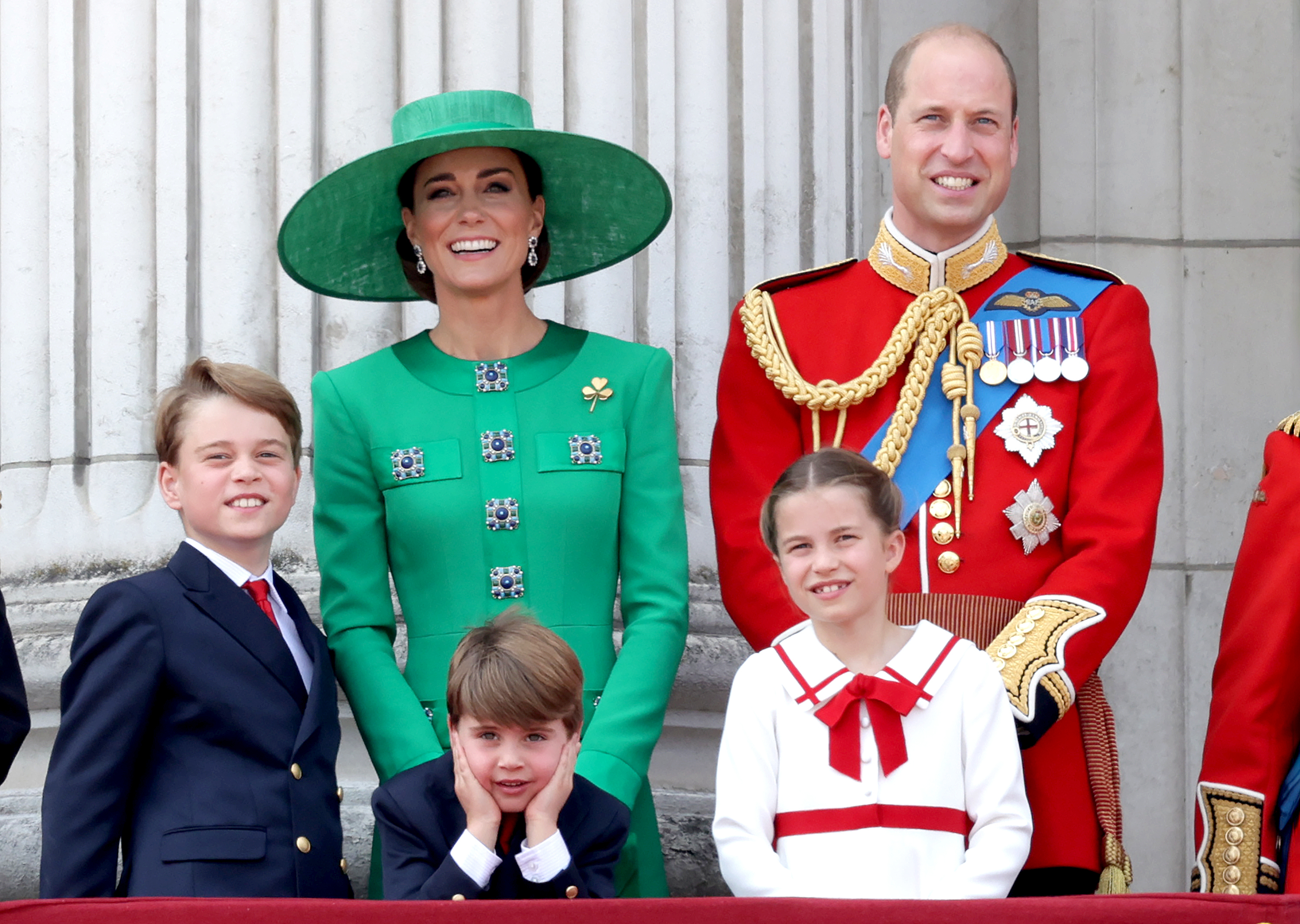 Royal family in formal attire on a balcony, two adults in ceremonial dress, two boys in suits, girl in dress with red accents