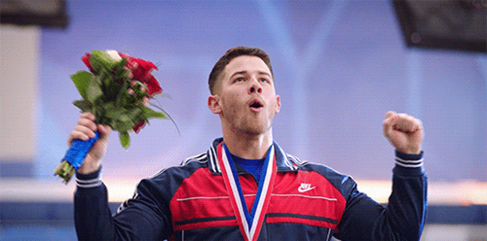 gif of joe jonas celebrating with a bouquet and a medal, wearing a sports jacket with a zipper