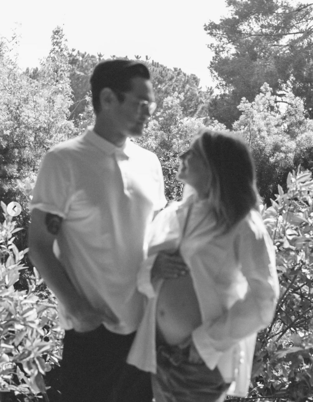 Christopher in a buttoned shirt and Ashley  in an open shirt cradling her visible baby bump, both smiling, with trees in the background