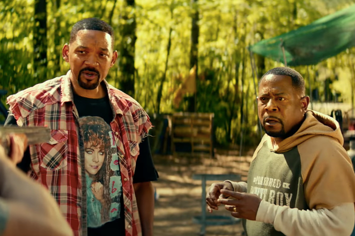 Two actors in a forest scene, one in a flannel shirt, the other in a hoodie and jacket. They appear in a tense conversation