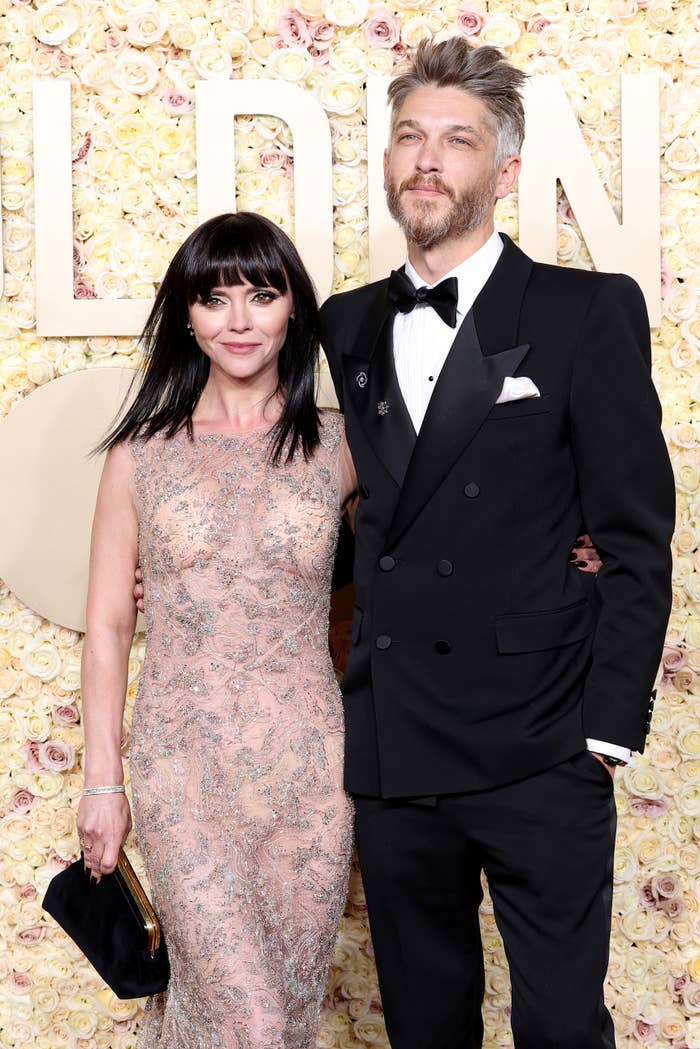 Christina in a sleeveless lace dress, holding a clutch, and Mark in a black tuxedo at a media event