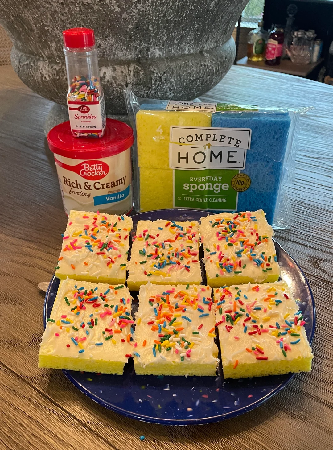 Plate of frosted cake squares with sprinkles; background shows a bottle of sprinkles, a sponge pack, and a frosting container