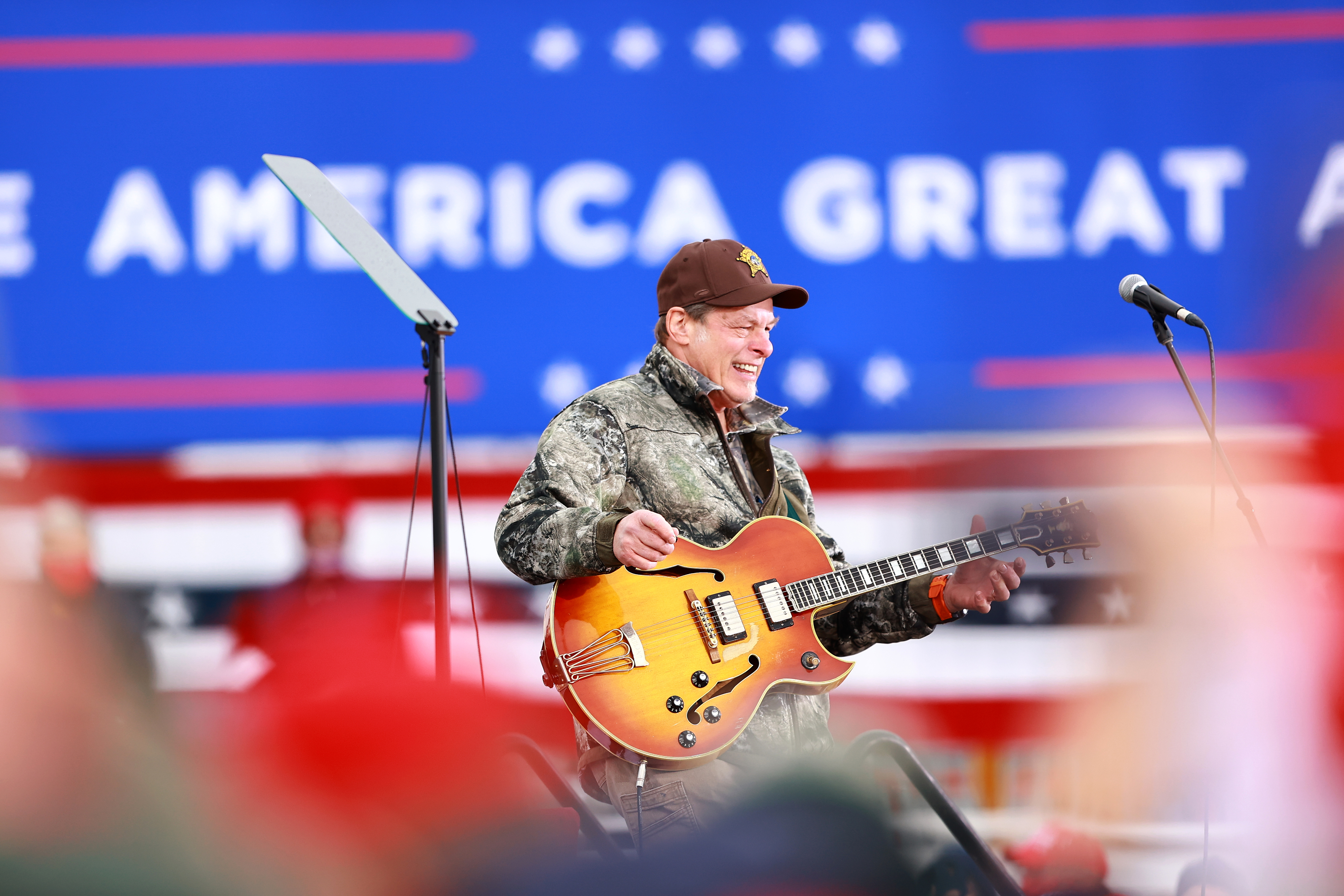 Ted in camouflage jacket playing an electric guitar at a rally with &quot;Make America Great Again&quot; sign in background