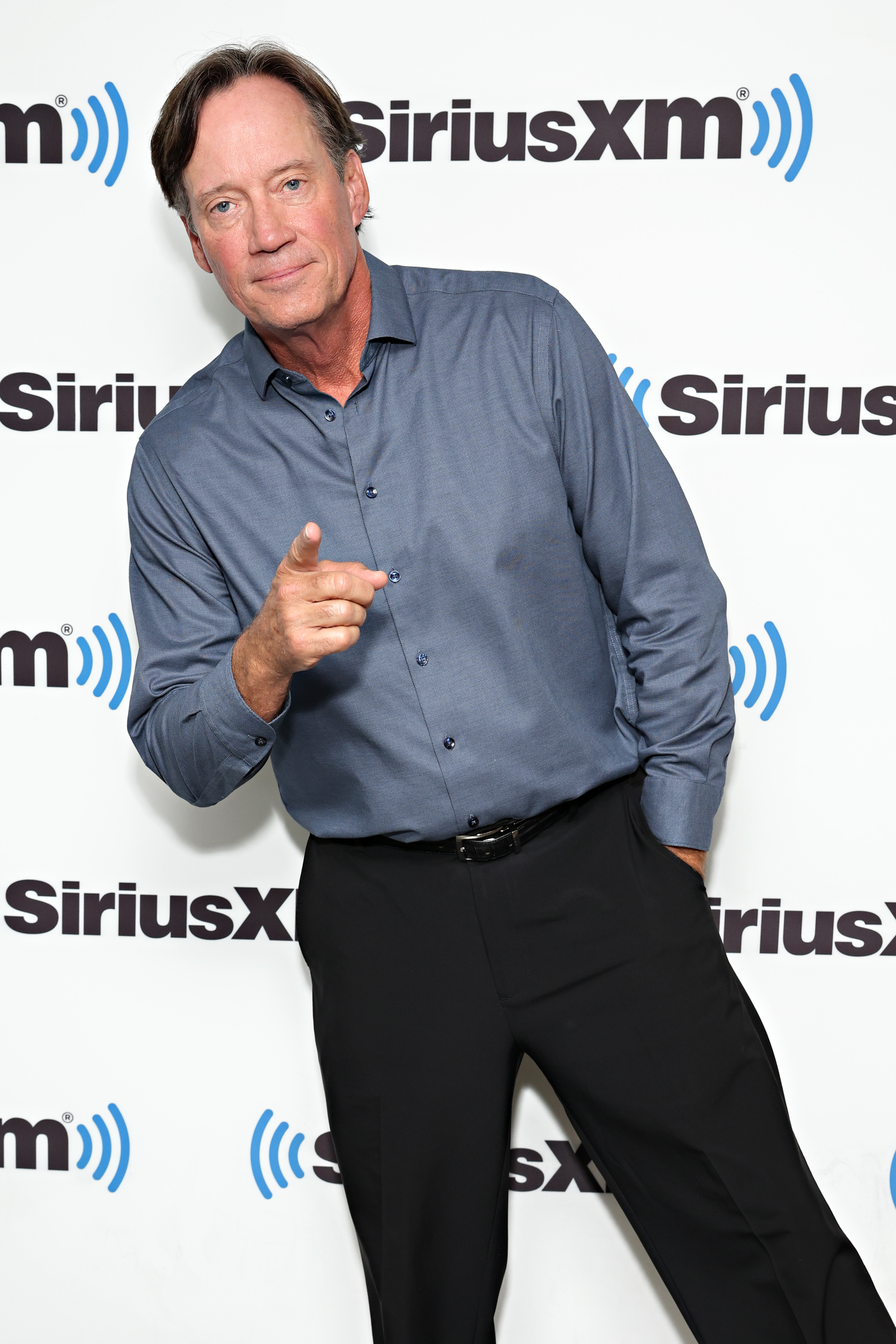 Kevin Sorbo in a blue shirt and black pants, pointing toward the camera at a SiriusXM event