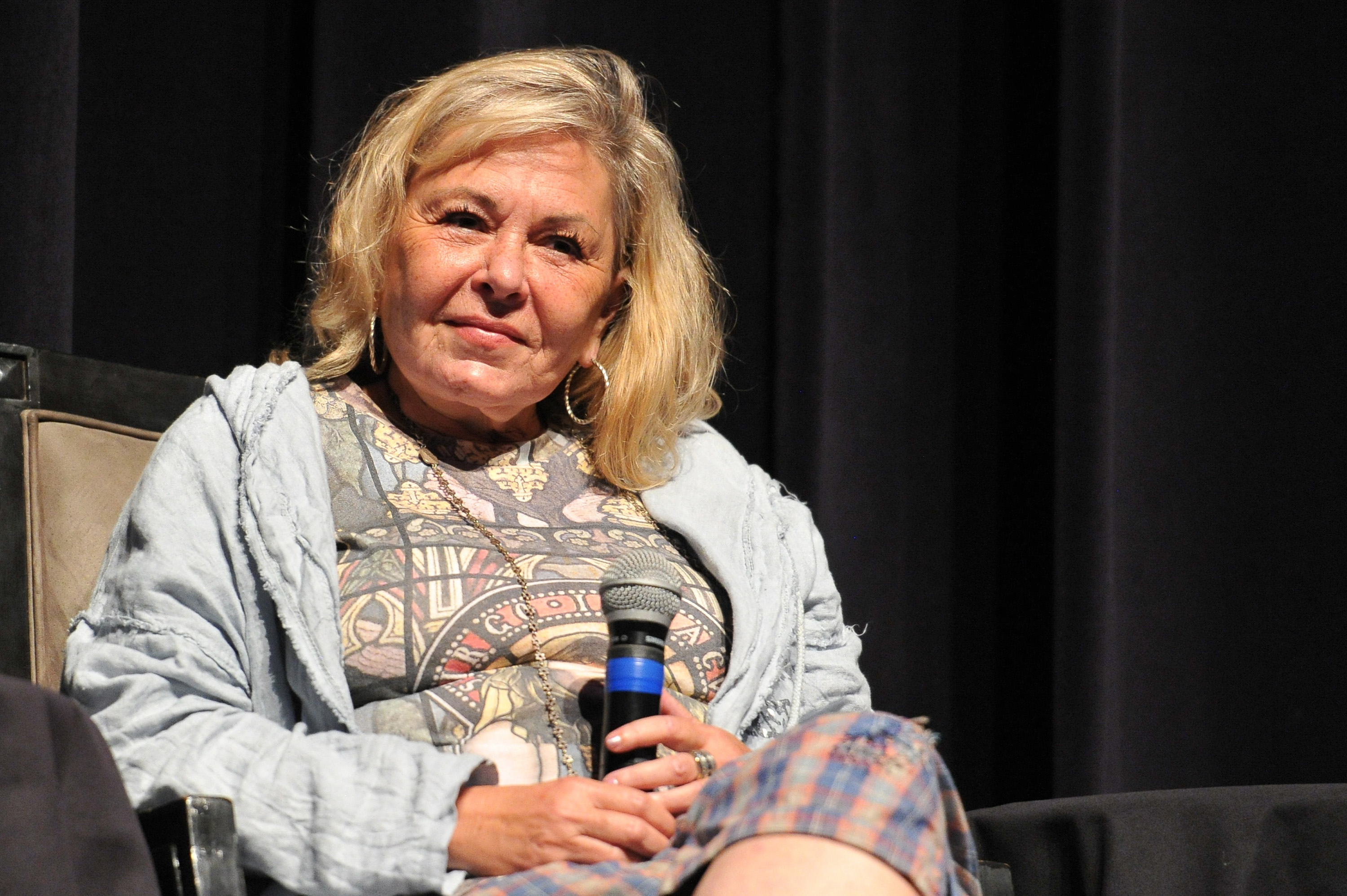 Roseanne sitting and holding a microphone, wearing a patterned shirt and a gray hoodie